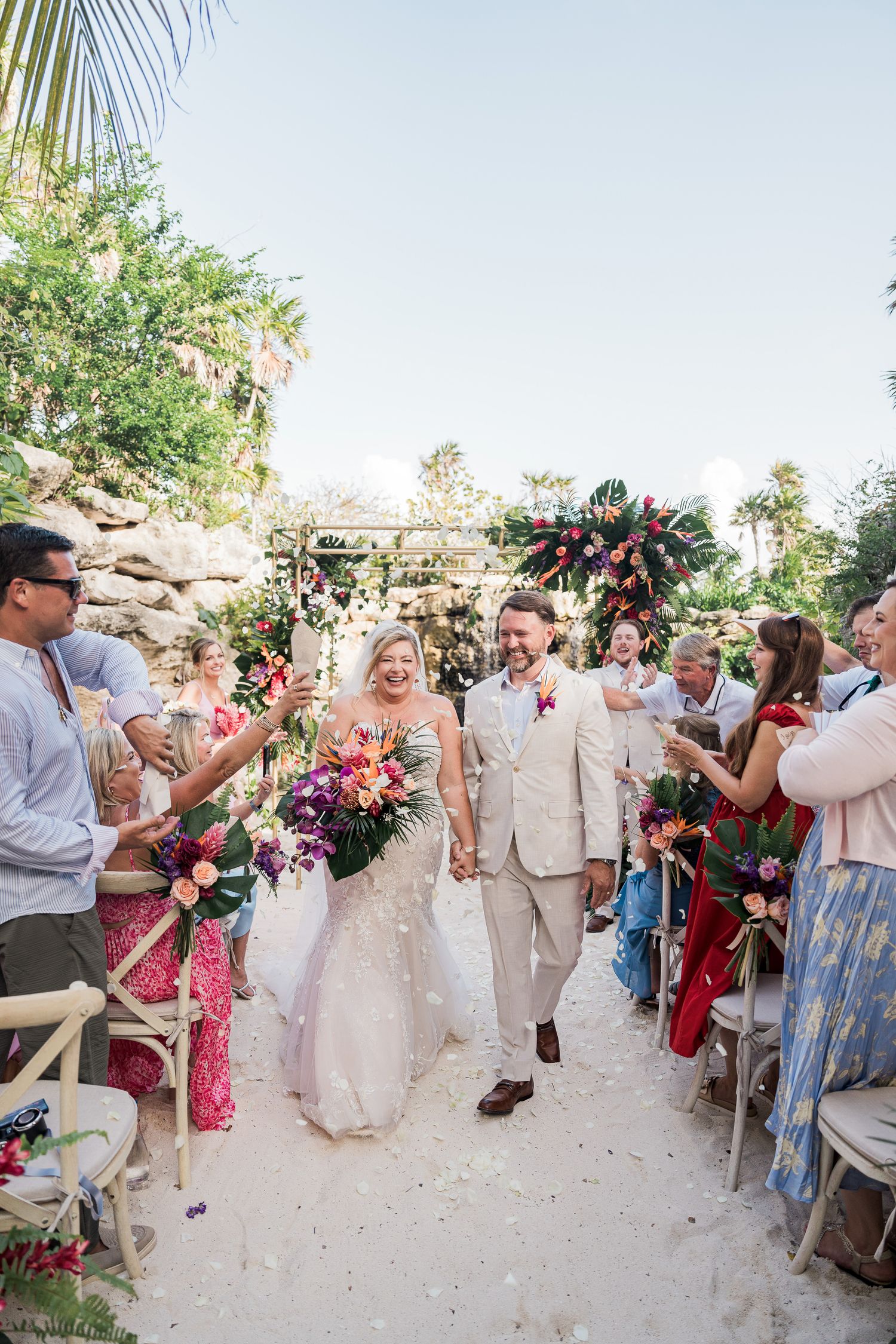 Bride and groom walk down sandy aisle as guests toss petals at tropical outdoor wedding ceremony with colorful floral arch.