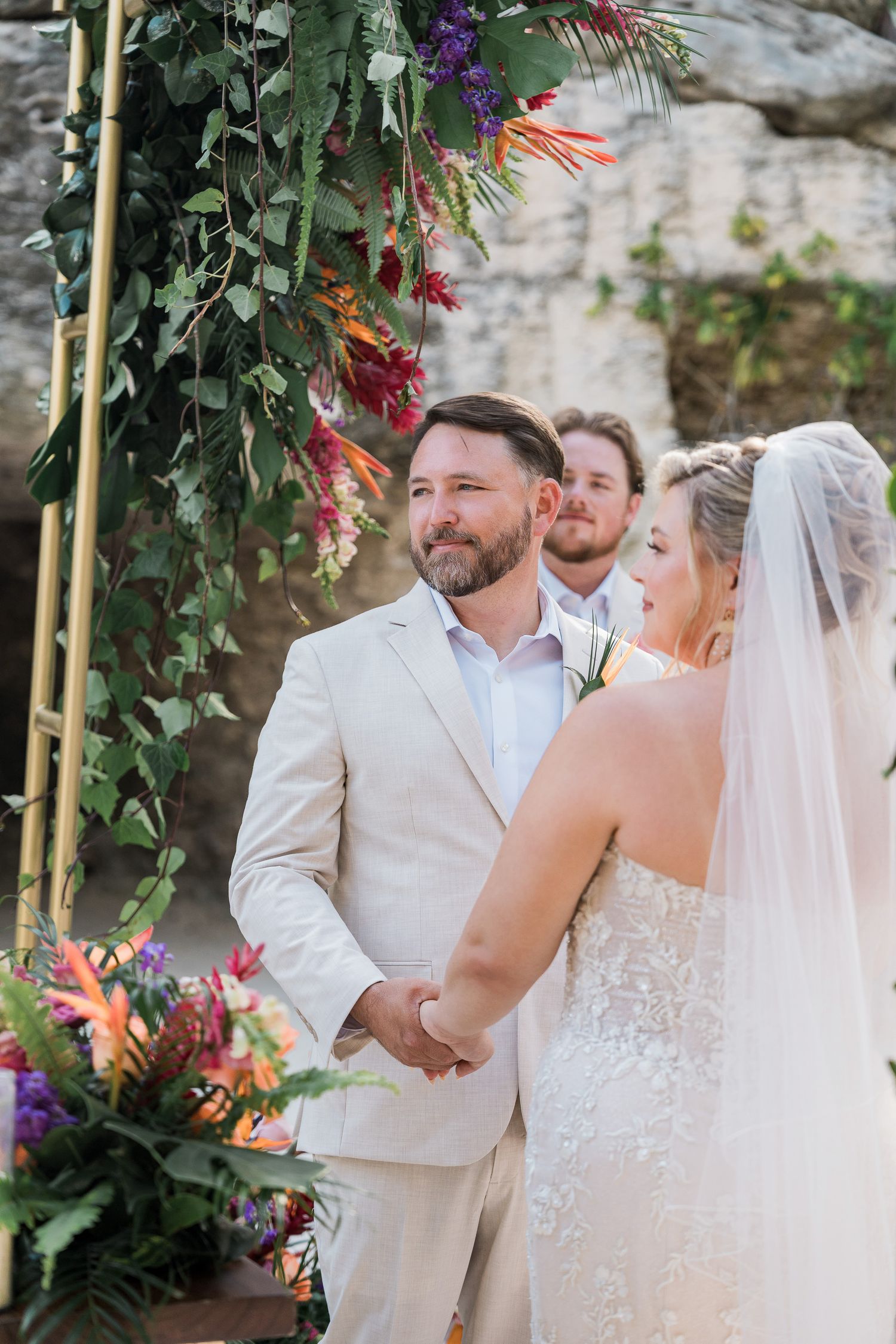 Bride and groom holding hands at outdoor wedding ceremony with colorful floral arch and stone backdrop.