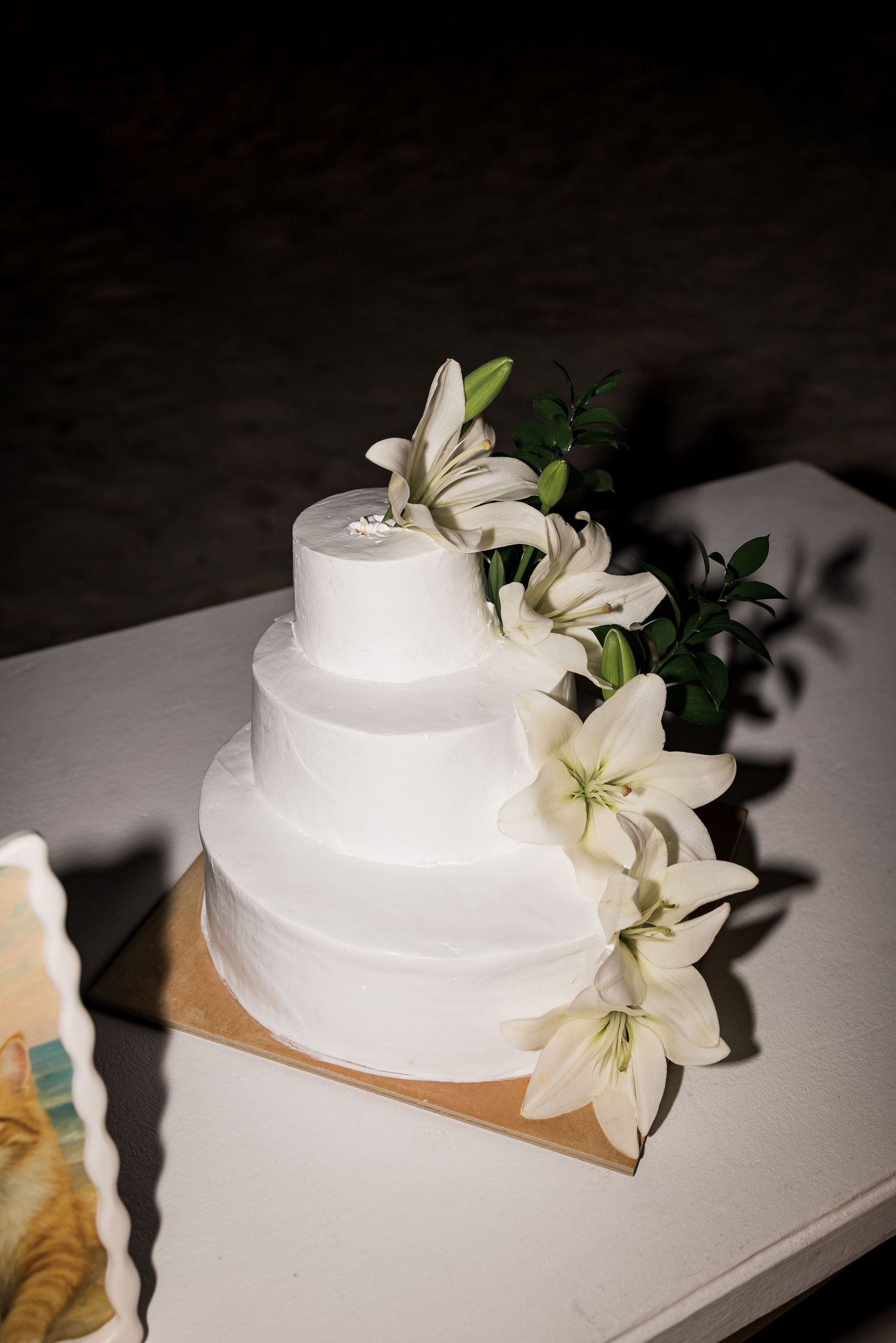 Elegant three-tier white wedding cake decorated with fresh white lilies and green foliage on a kraft board.