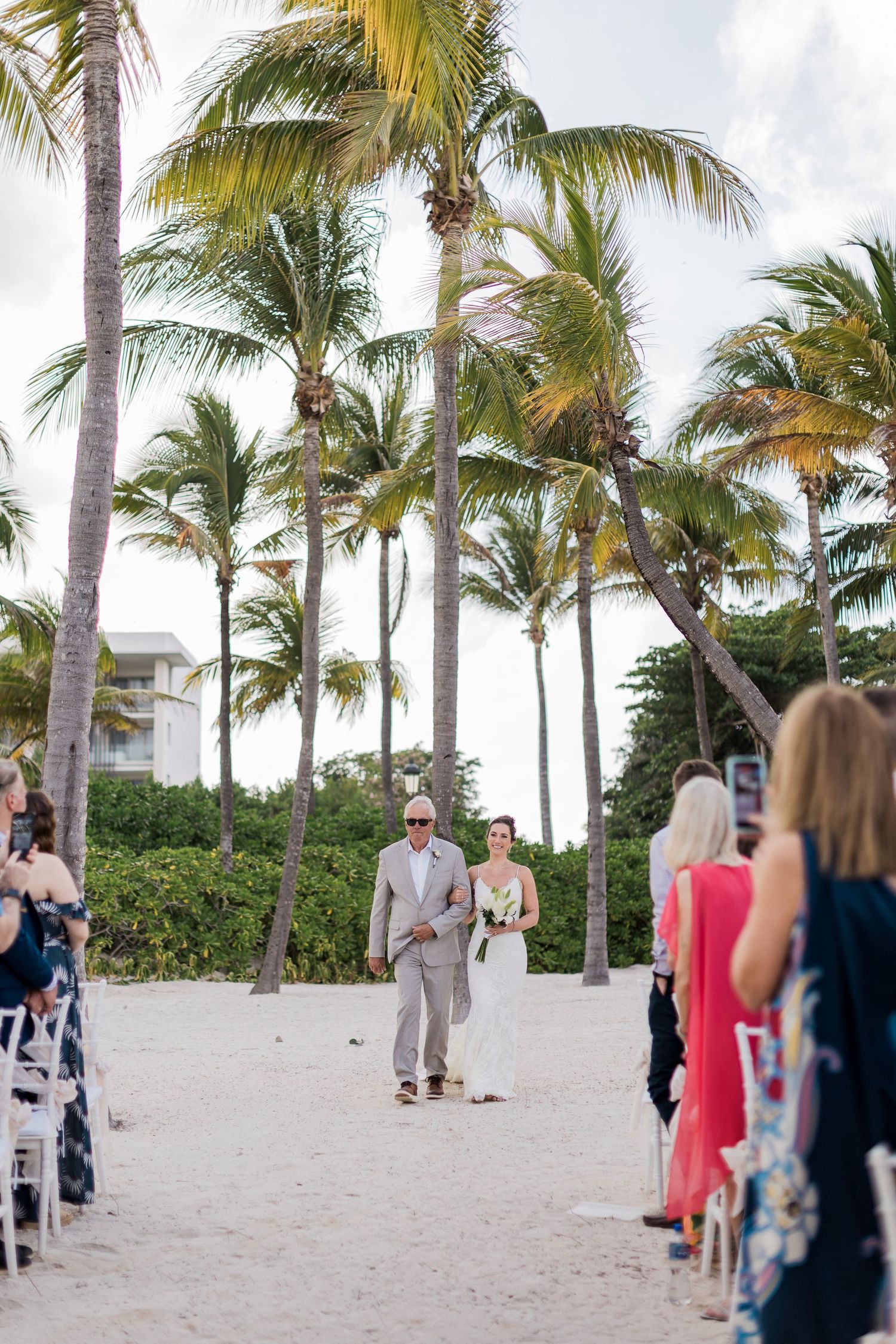 Bride walks down sandy beach aisle with escort among palm trees at tropical outdoor wedding ceremony.