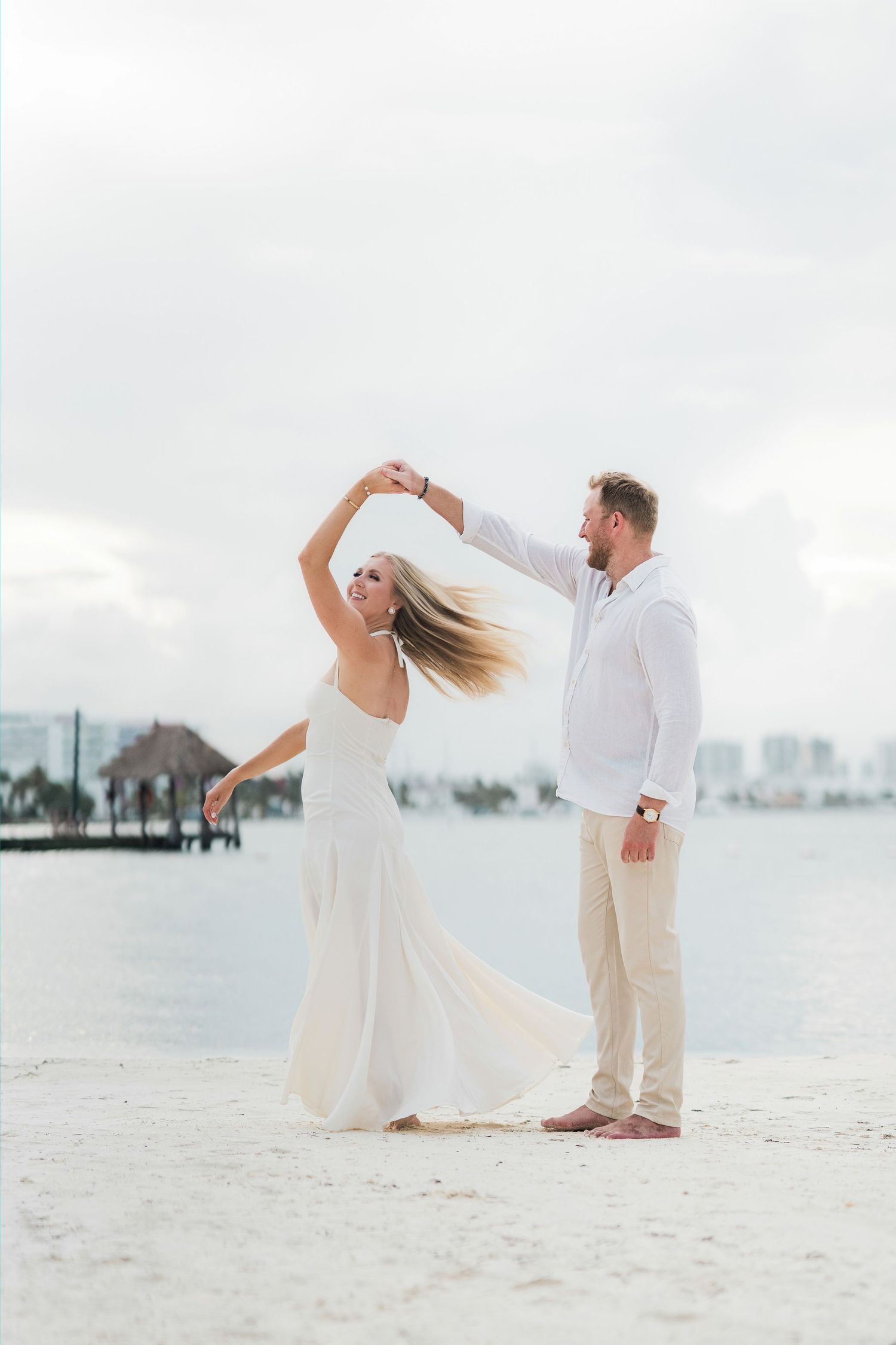 Cancun Engagement Photoshoot - Couple dancing on a white sand beach in white outfits, man spinning woman near tropical waterfront.