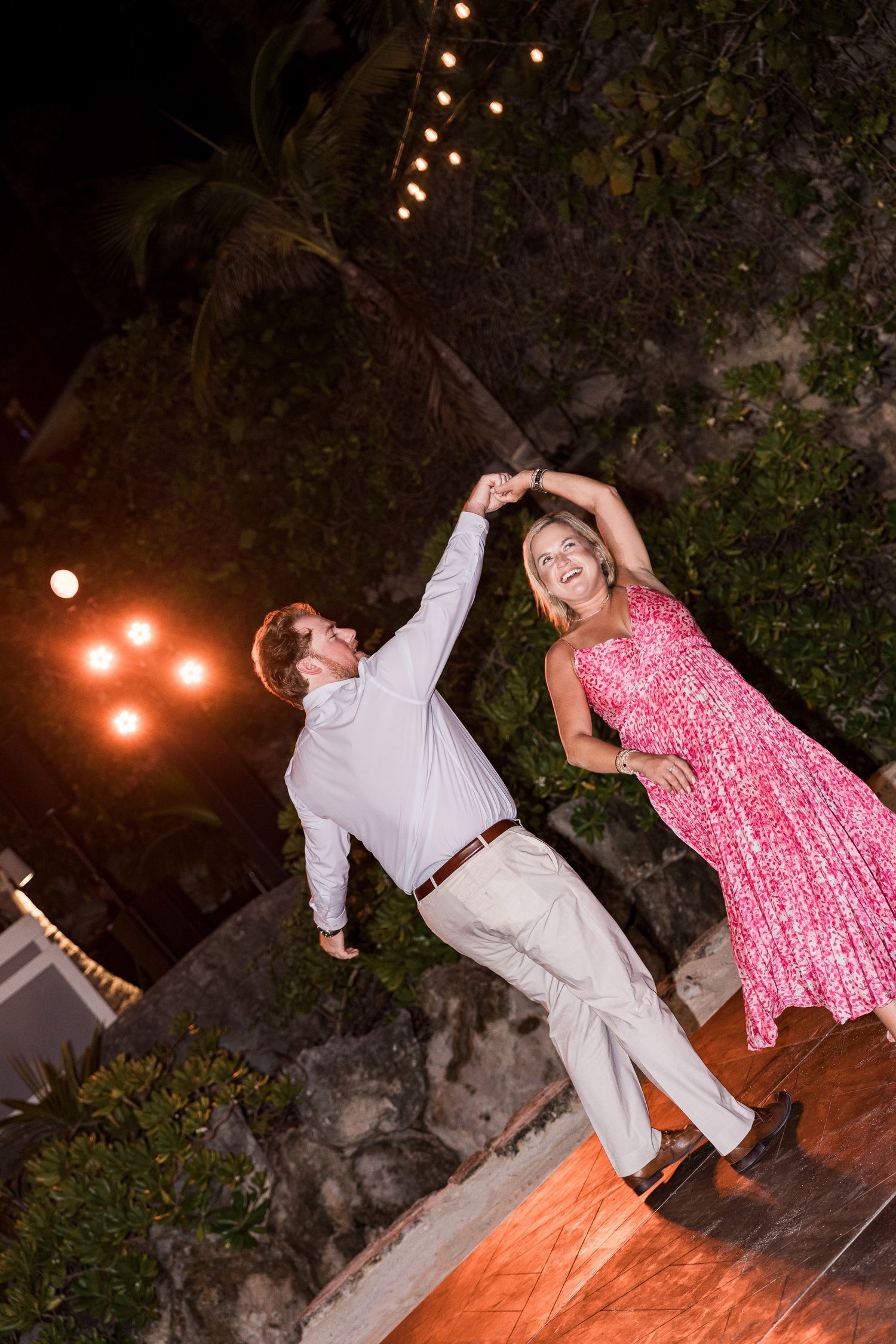 Couple dancing outdoors at night under string lights, woman in pink floral dress spinning with man in white shirt.