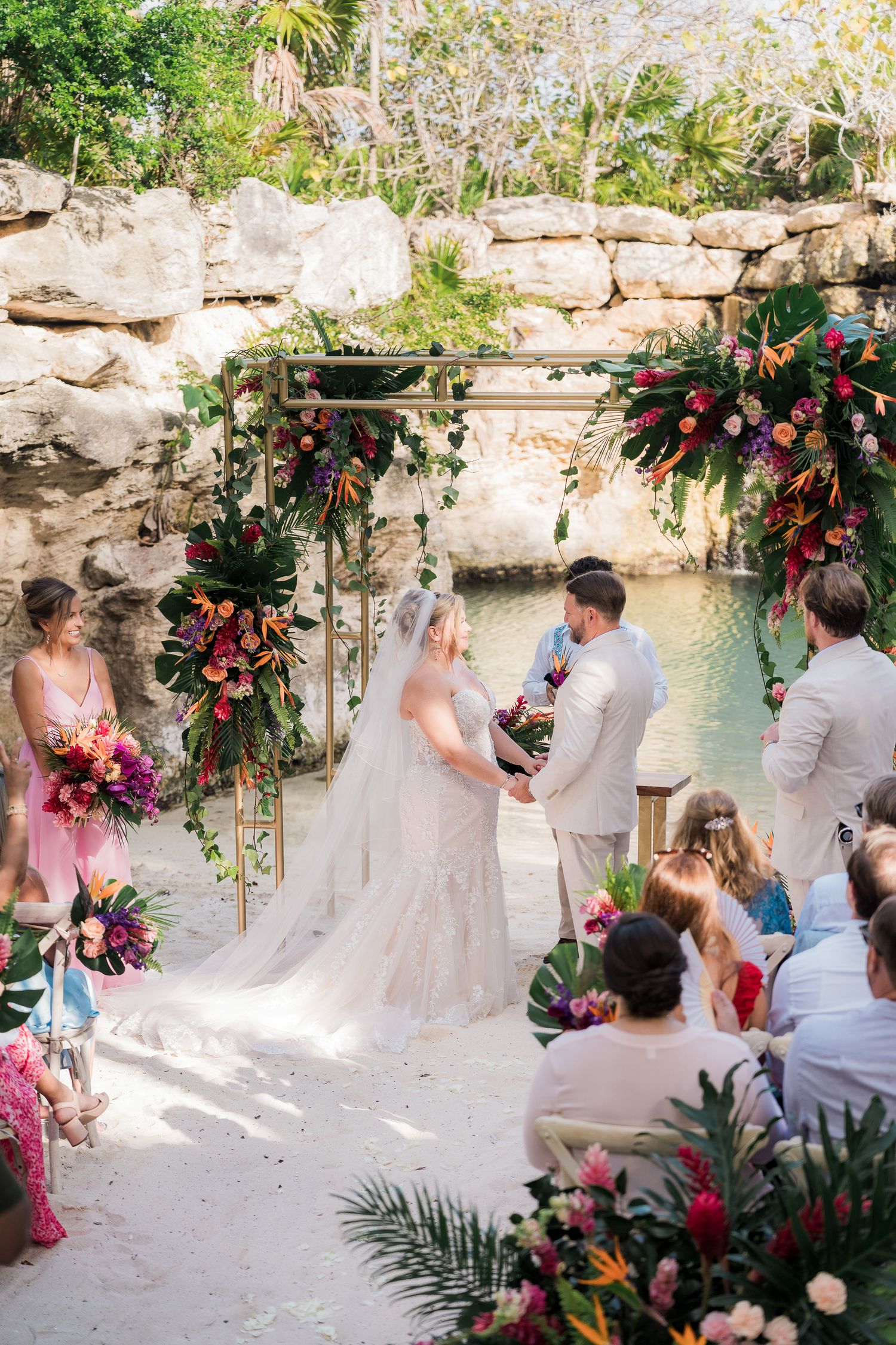 Hotel Xcaret México Tropical beach wedding ceremony with bride and groom exchanging vows under floral arch  by turquoise water.