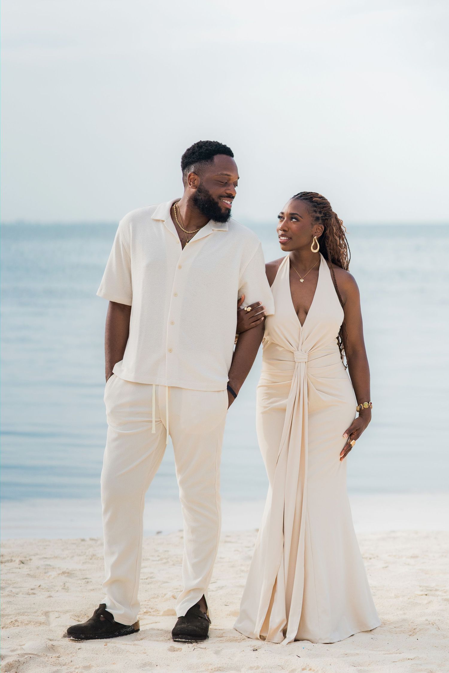 Elegant couple in coordinating ivory outfits smiling at each other while strolling on a sunny beach.