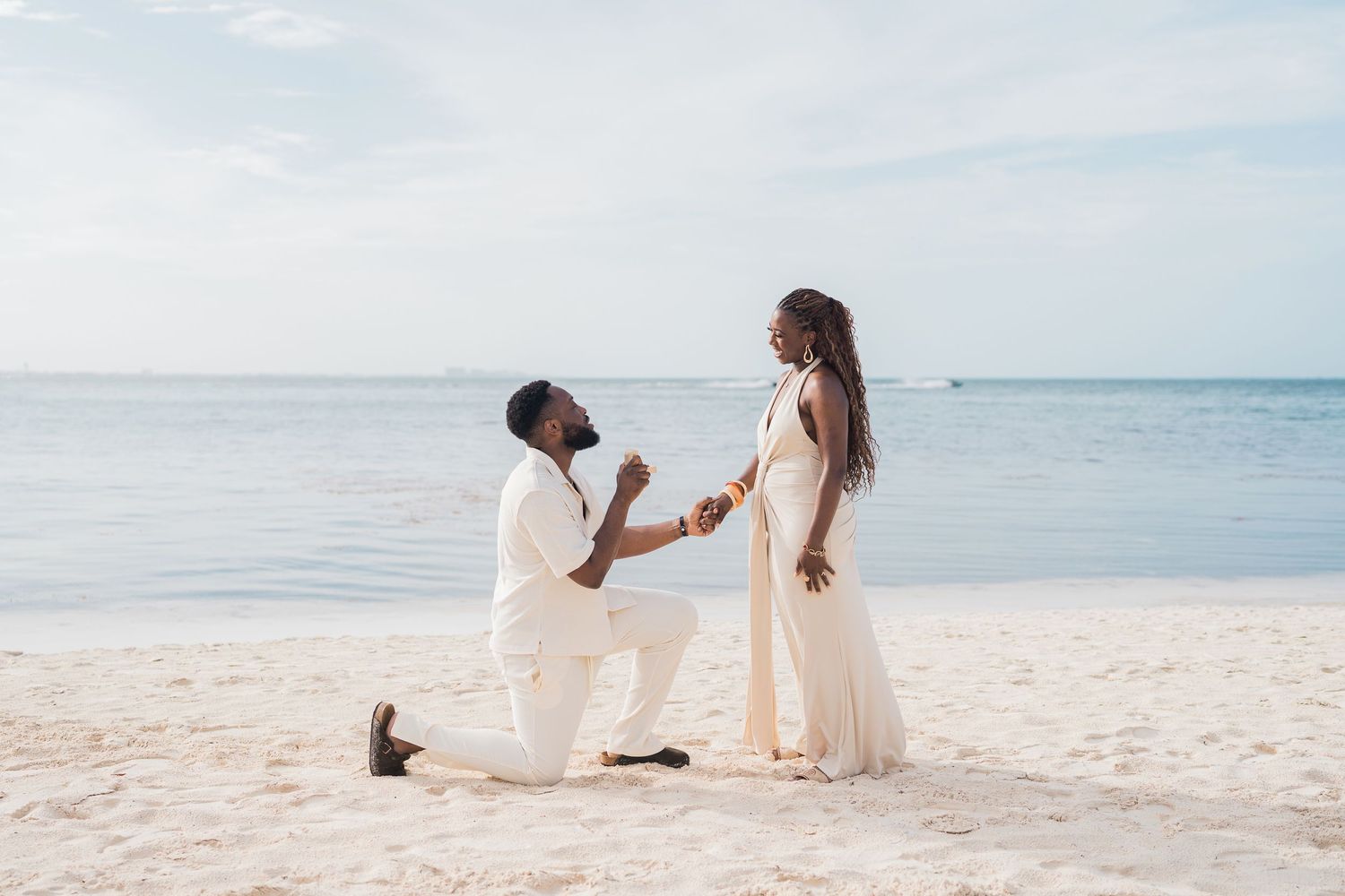 Man proposing to woman on a sandy beach, both dressed in white, with calm ocean waters in the background.