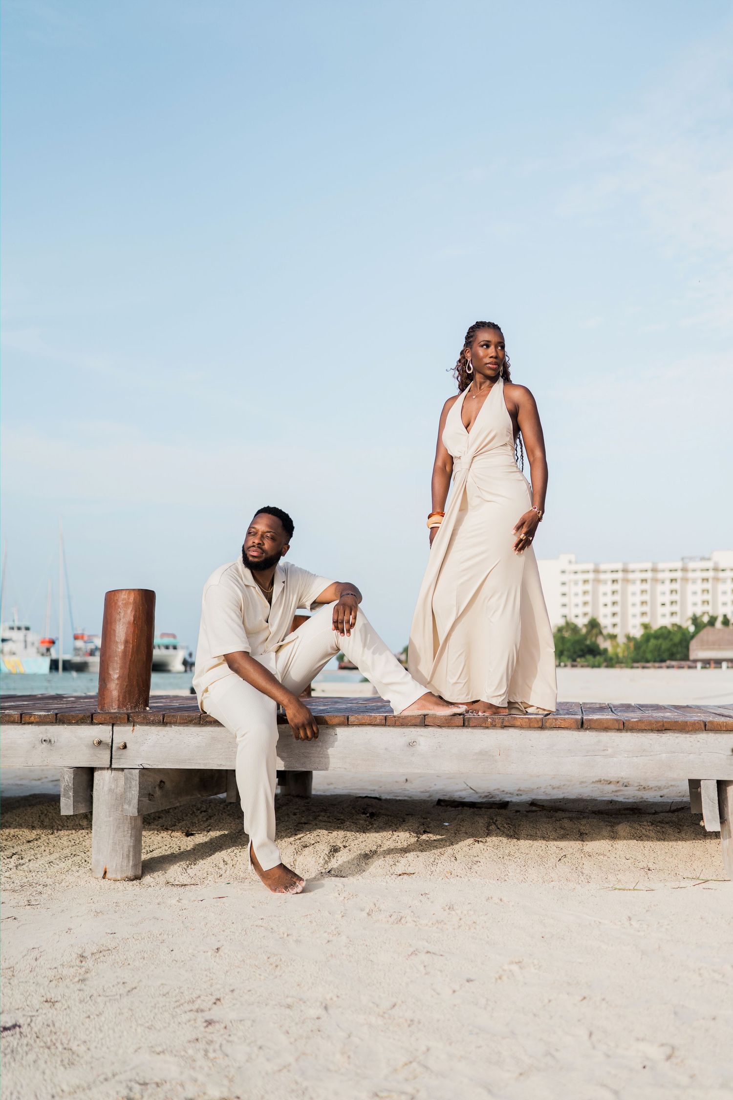 Couple in matching cream outfits posing on a beachside wooden bench with a coastal backdrop.