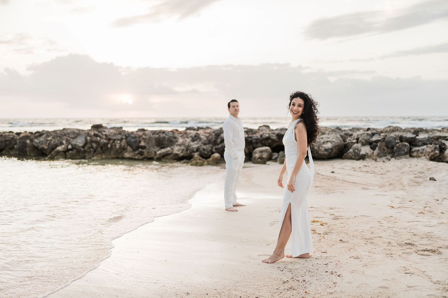 Couple in white outfits posing on a sandy beach at sunset with rocky shoreline in the background.