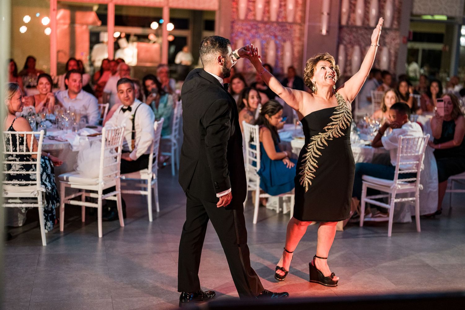 Woman in black dress raises arm while dancing with man in tuxedo at lively wedding reception.
