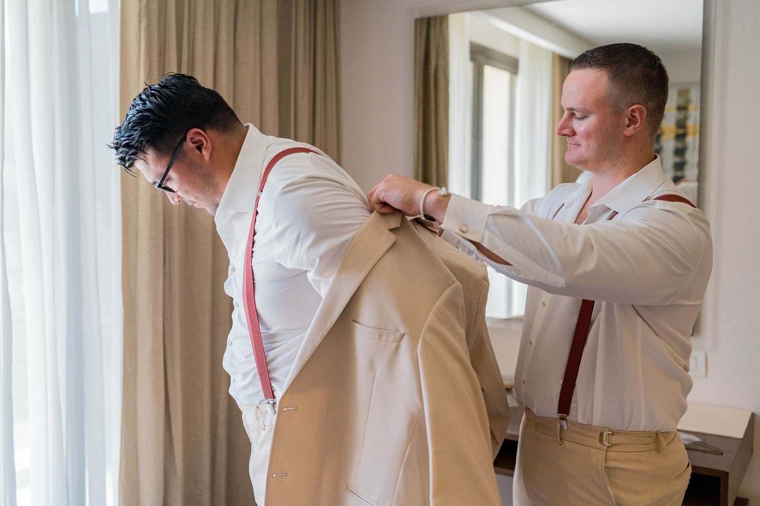 Groomsman helping groom put on a beige suit jacket with red suspenders before the wedding ceremony.