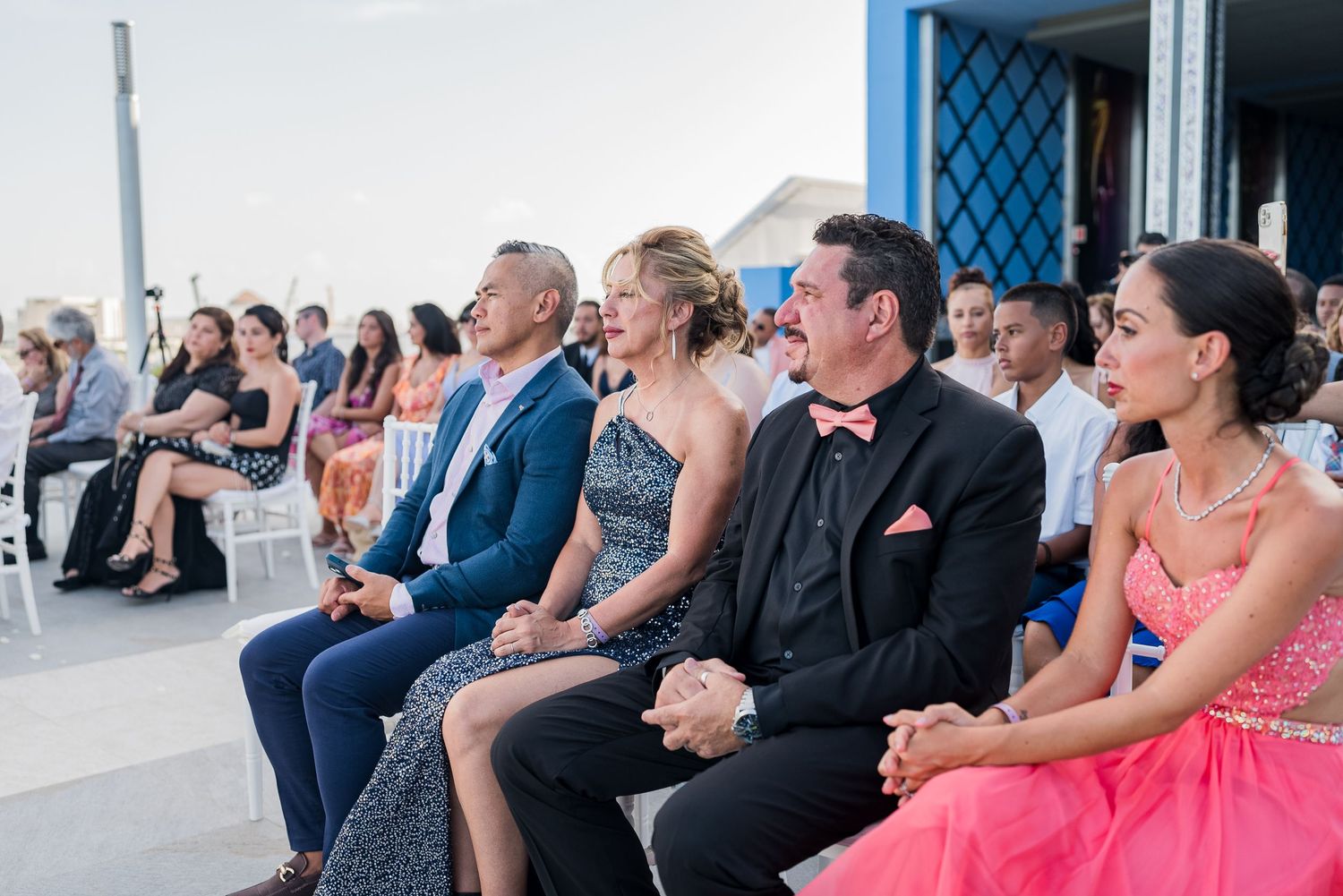 Wedding guests seated outdoors watching ceremony in colorful attire at beachside venue.