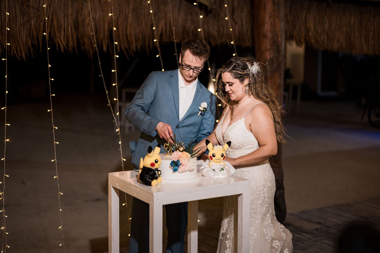 Bride and groom cut whimsical Pokemon-themed wedding cake under fairy lights at reception.