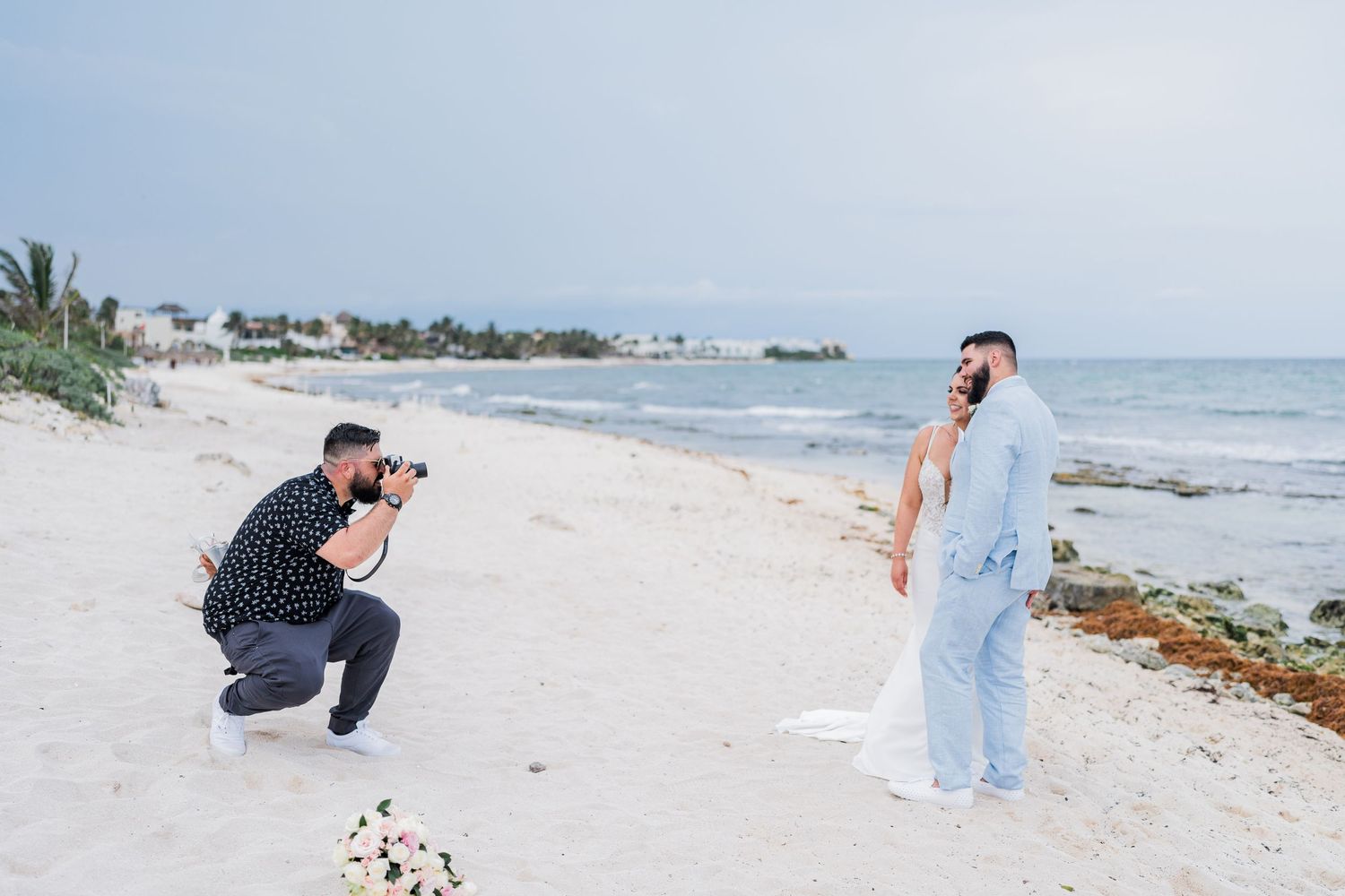 Wedding photographer kneeling on sandy beach to capture photos of bride and groom during outdoor beach photo session.