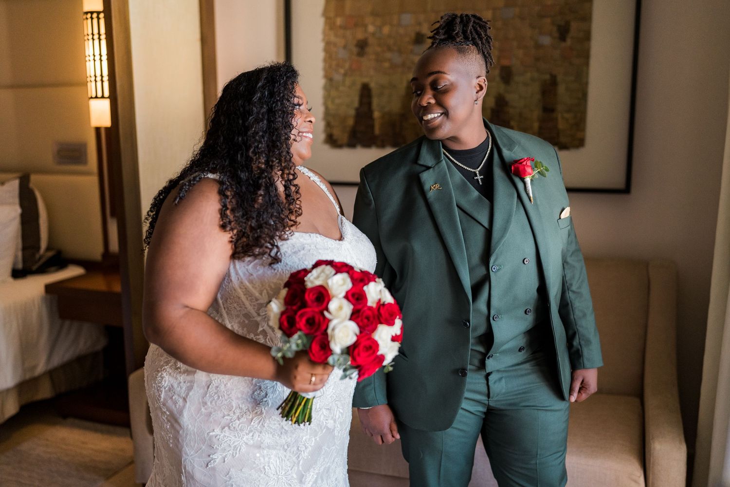 Lesbian couple sharing first look moment, one in white lace wedding gown holding red roses bouquet.