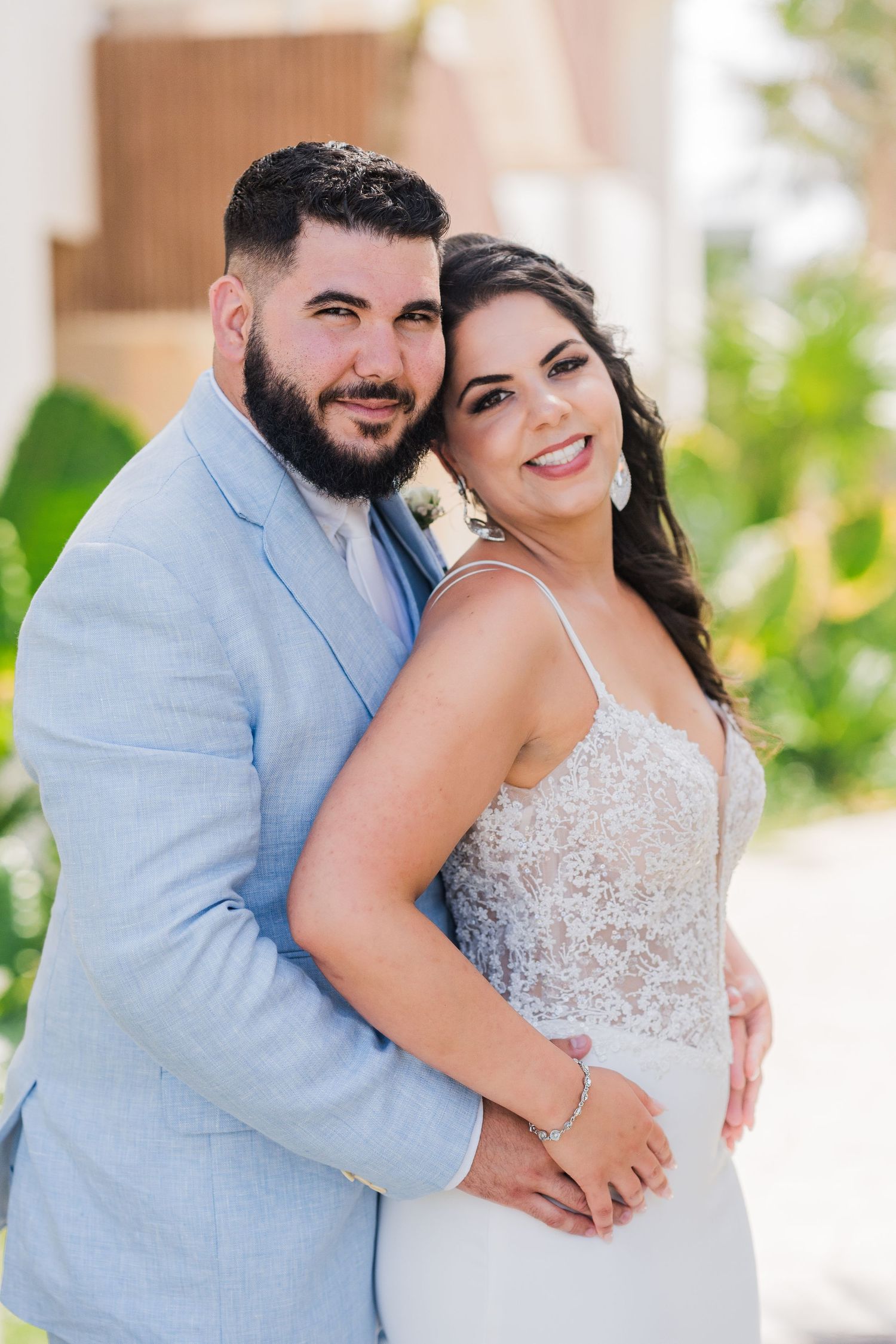 Groom in light blue suit embracing smiling bride in lace wedding gown at tropical outdoor venue.