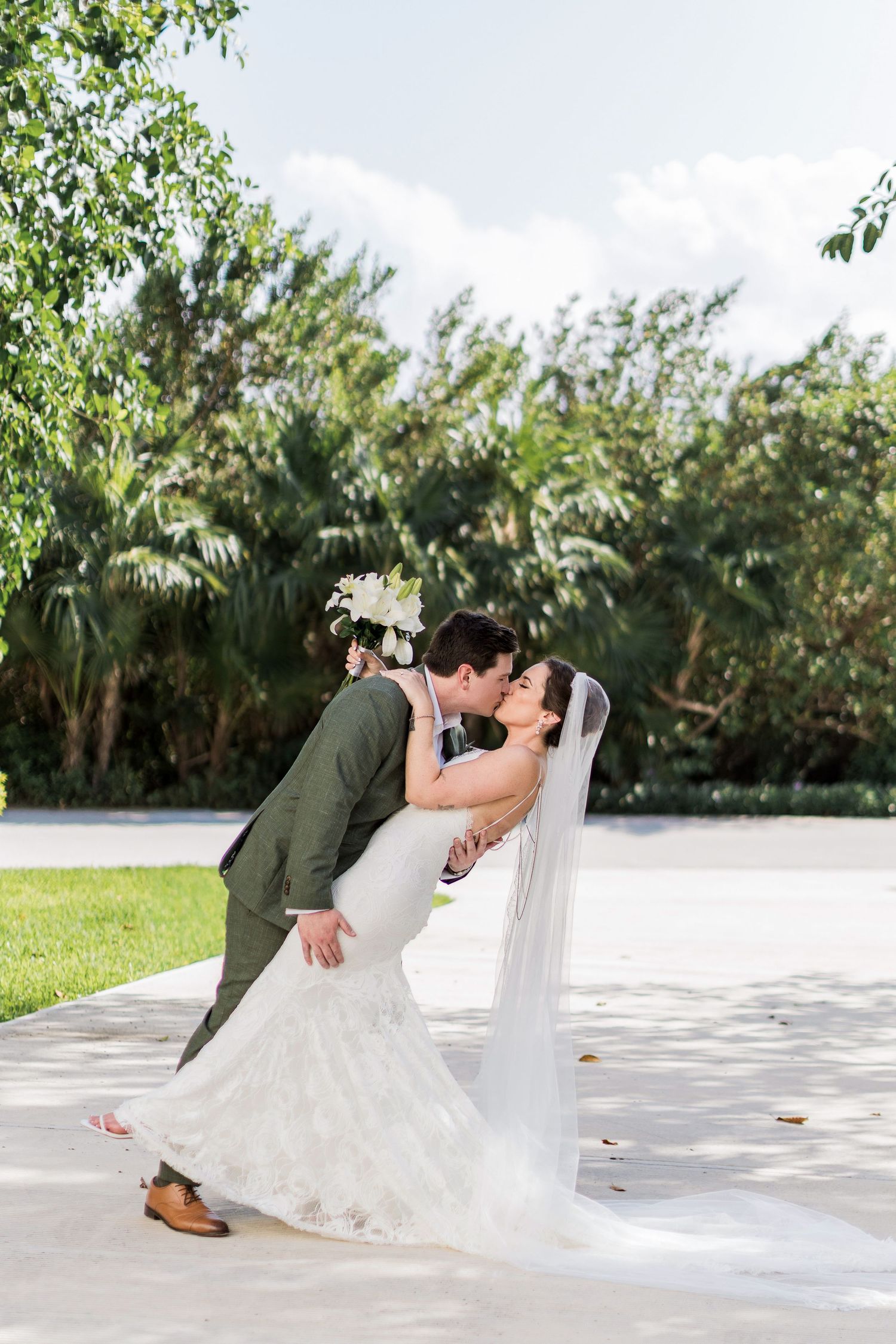 Bride and groom share a romantic dip kiss outdoors, with the bride in a lace gown and veil holding white flowers.