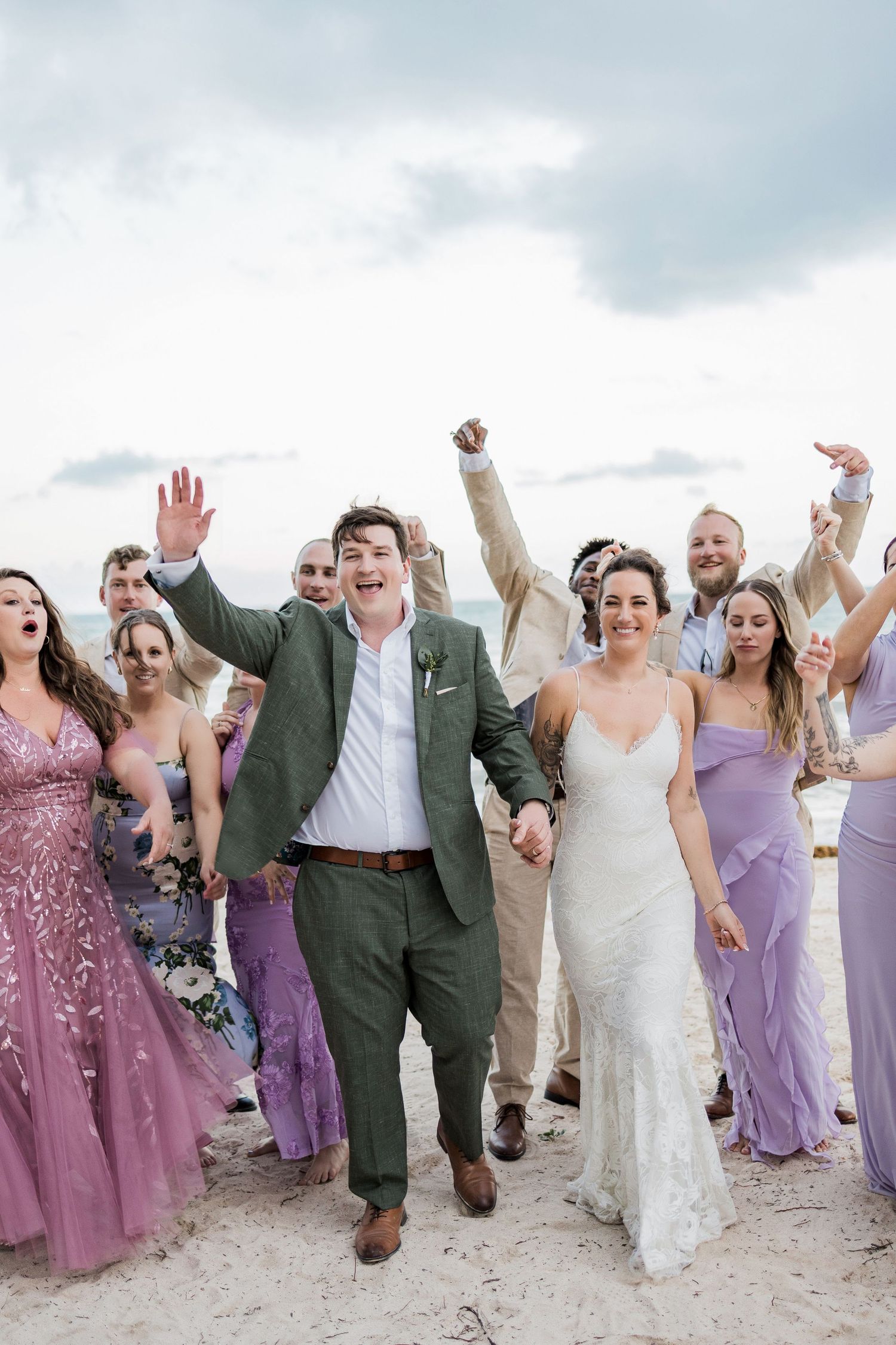 Joyful newlyweds hold hands on a beach, surrounded by cheering wedding party in lavender dresses and neutral suits.