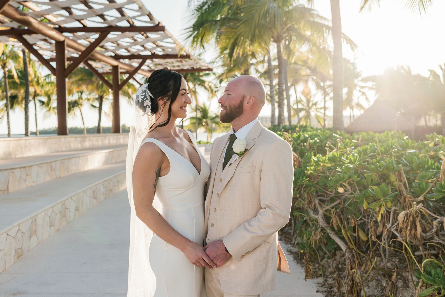 Bride and groom share a romantic moment on a tropical beach resort pathway, with palm trees and golden sunset light.
