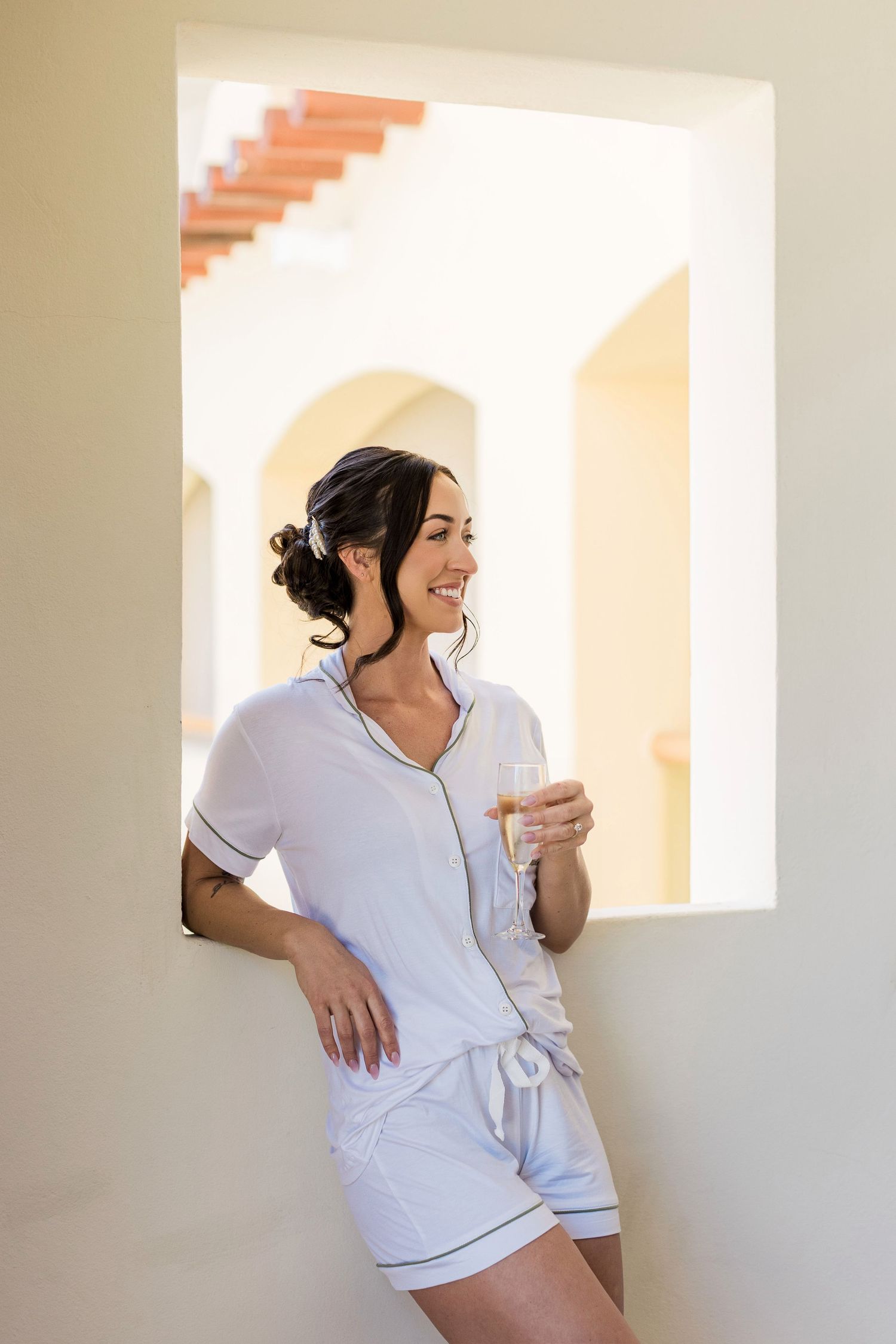 Woman in white pajama romper holding champagne glass, smiling on a bright resort balcony.