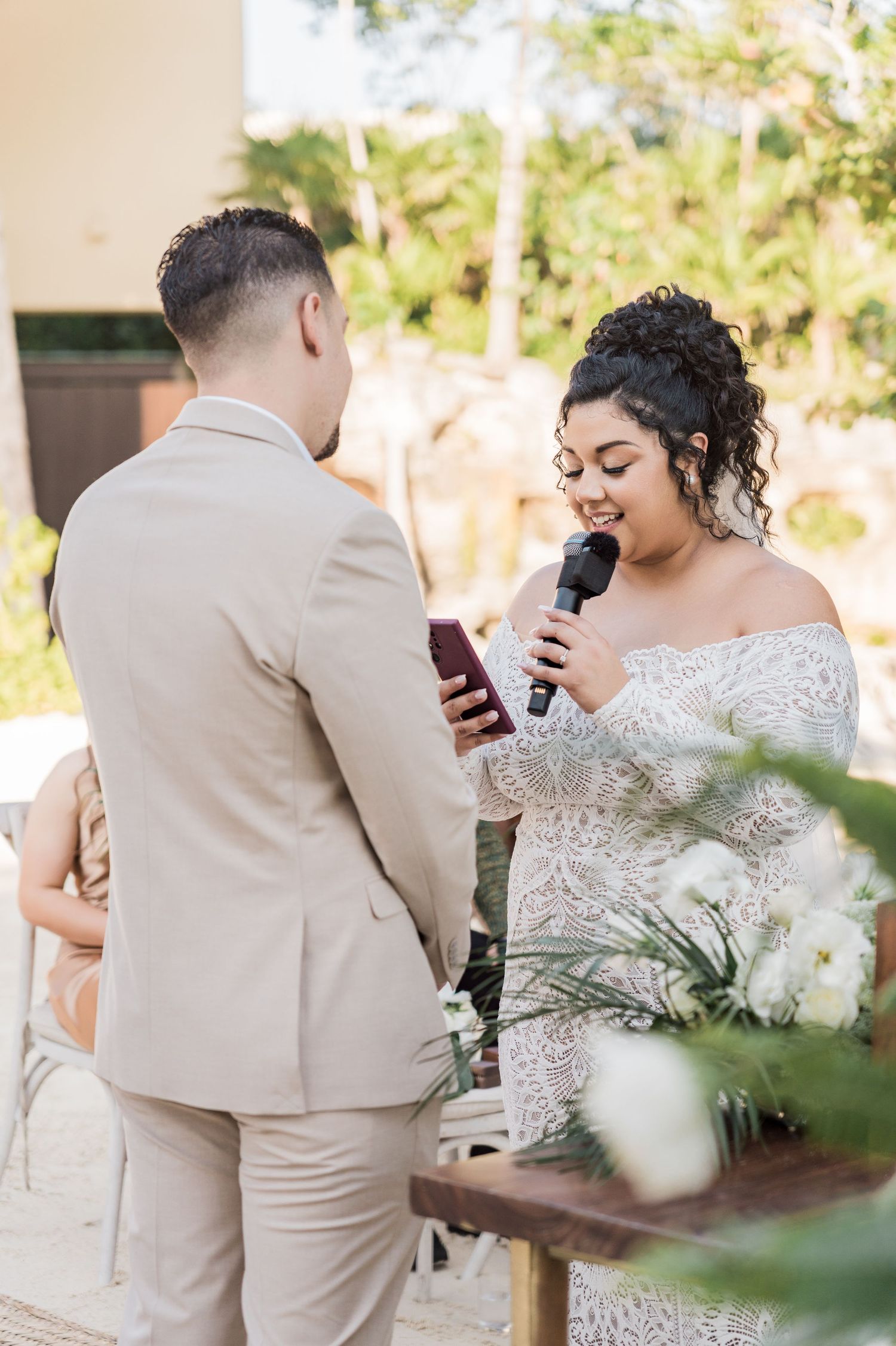 Xcaret Arte Wedding - Bride in lace off-shoulder gown reads vows from phone at outdoor wedding ceremony while groom listens in beige suit.