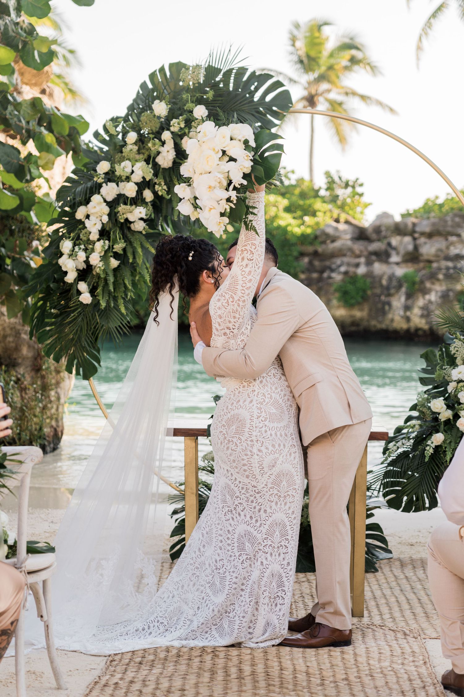 Xcaret Arte Wedding - Bride and groom share first kiss under floral arch with tropical greenery and white flowers