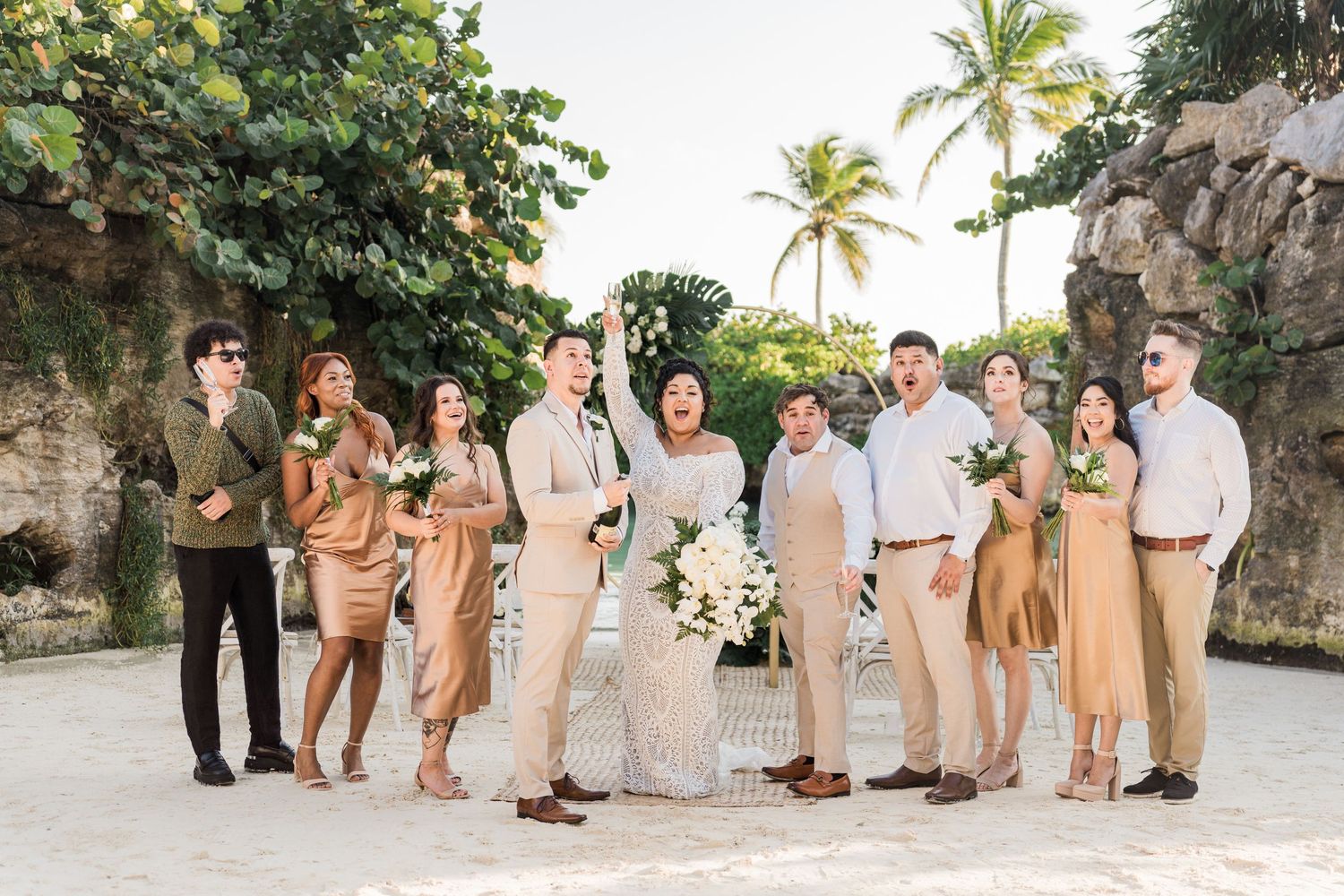 Xcaret Arte Wedding - Beach wedding party in neutral tones posing joyfully on white sand with palm trees and floral arch backdrop.