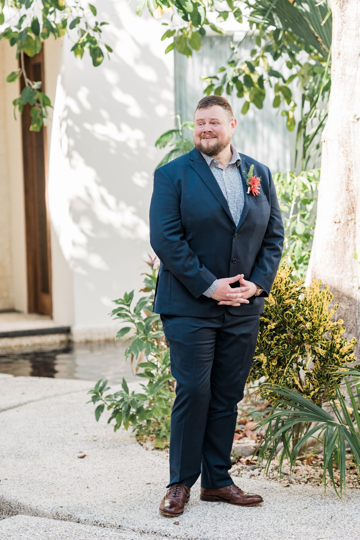 Secrets Akumal Wedding - Groom in navy blue suit with coral boutonniere smiling outdoors among lush greenery on wedding day.