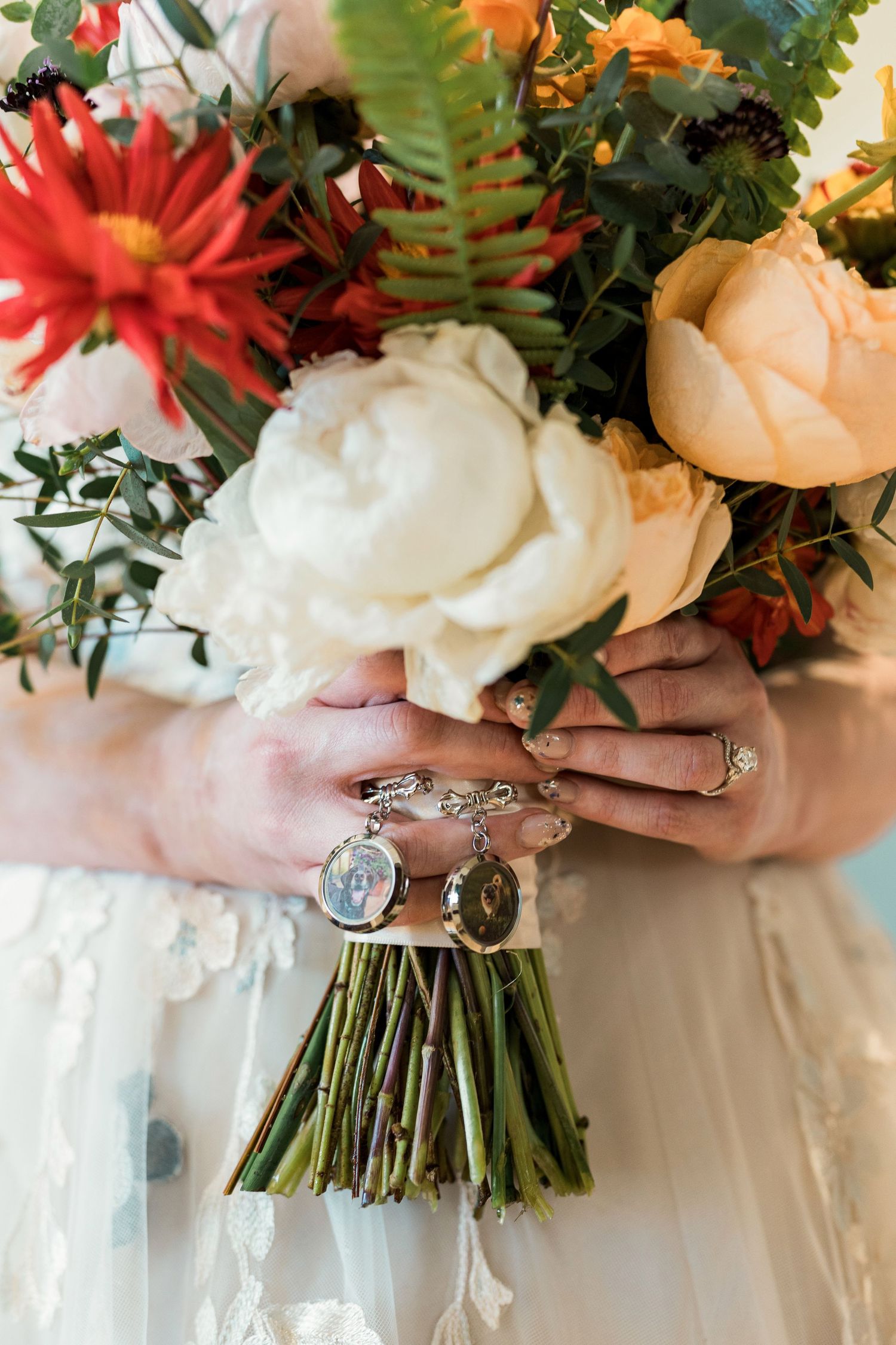 Bride holding a vibrant wedding bouquet with white and peach peonies, red dahlias, and locket charms tied to the stems.