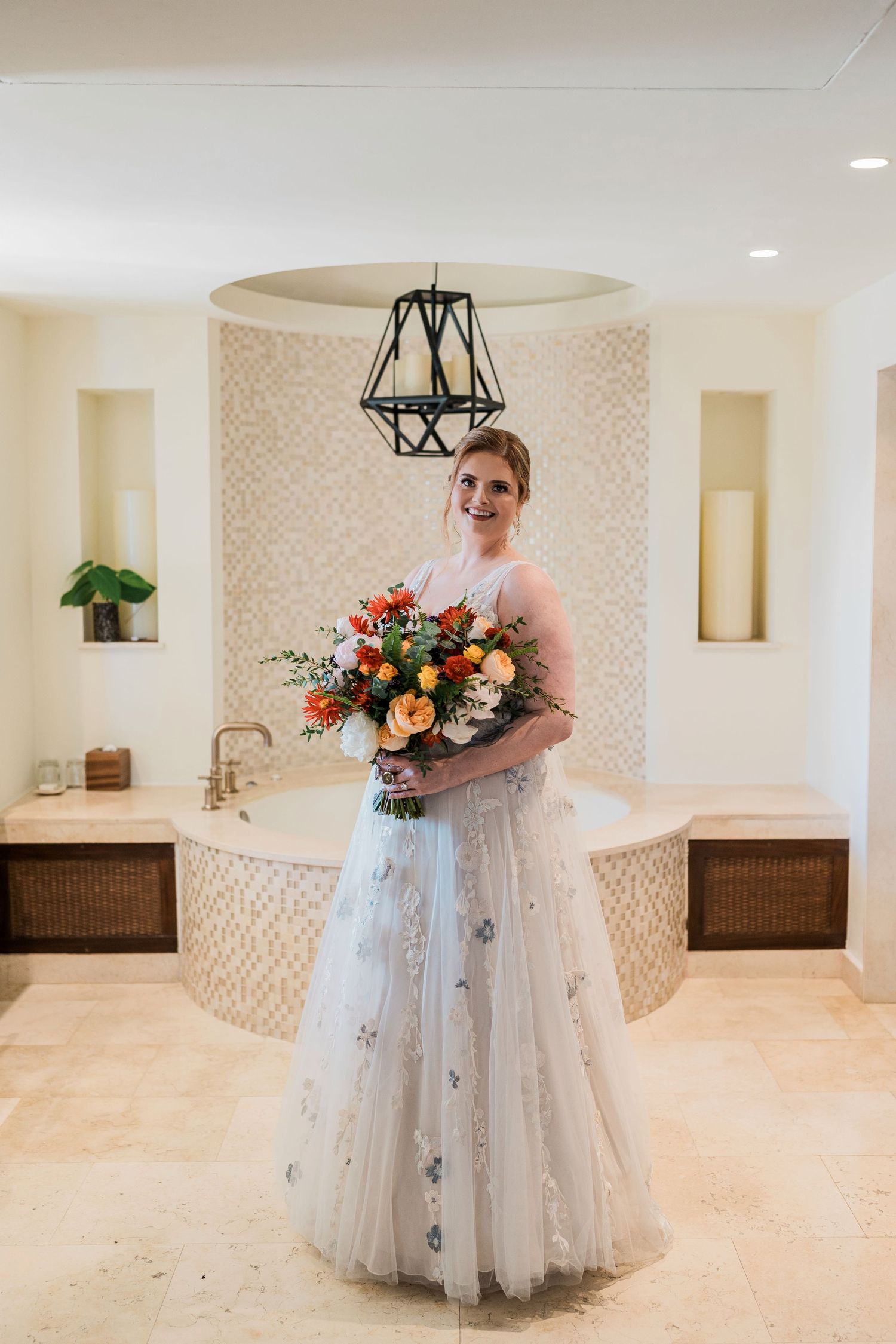 Smiling bride in floral tulle gown holding vibrant orange bouquet stands in elegant marble bathroom.