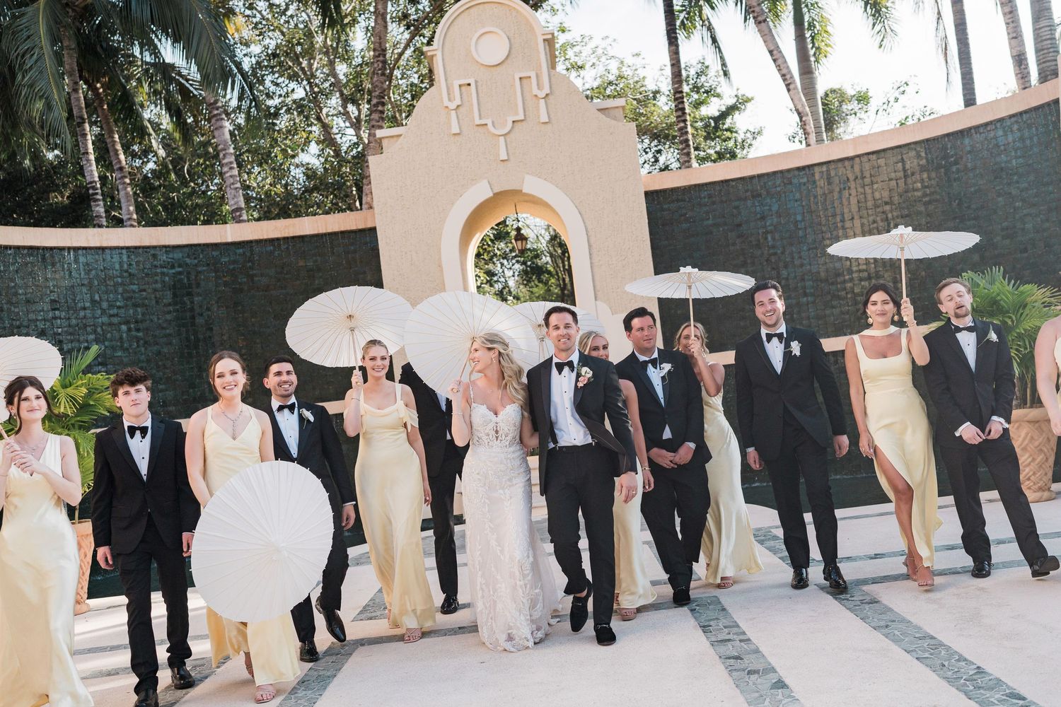 Wedding party poses outdoors in formal attire, bridesmaids in gold dresses holding white parasols, groomsmen in tuxedos.