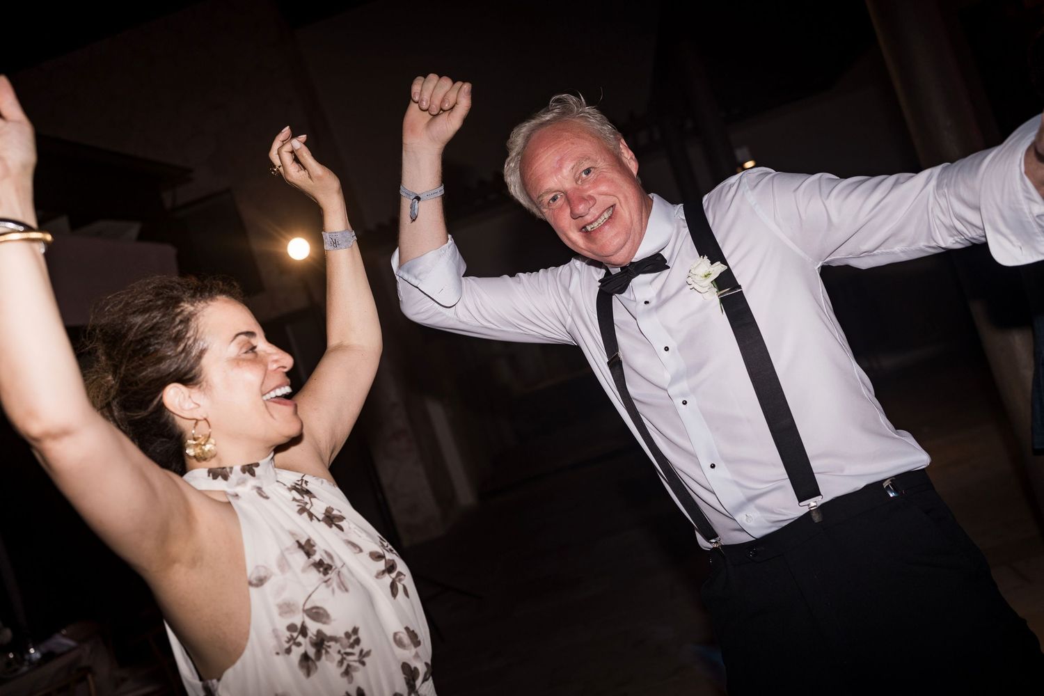 Two people dancing joyfully at a wedding reception, arms raised, woman in floral dress and man in white shirt with suspenders. TRS Yucatán