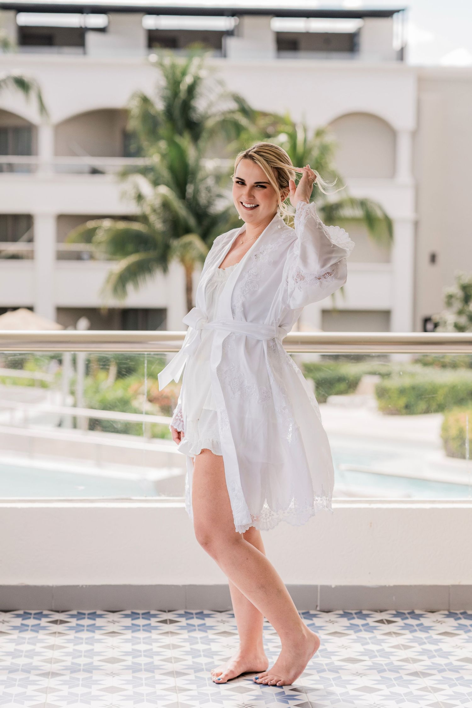 Smiling blonde woman in white robe standing barefoot on a resort balcony with palm trees and pool in background.