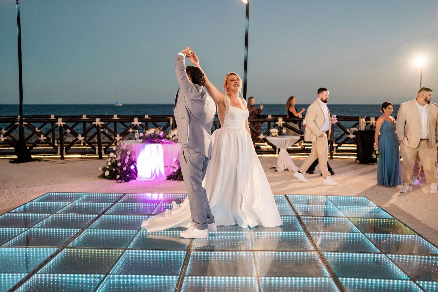 Bride and groom share first dance on illuminated LED dance floor at outdoor beach wedding reception at dusk.