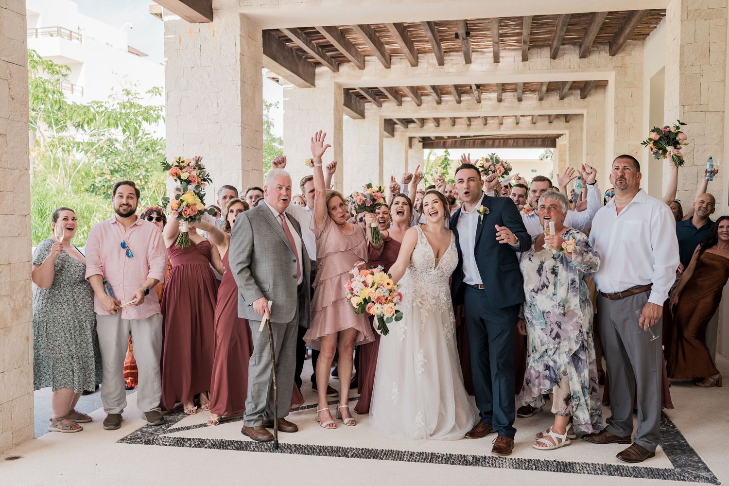 Joyful wedding party celebrating with raised hands in an elegant outdoor venue, bride holding colorful bouquet.