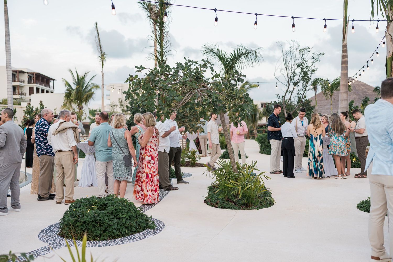 Guests mingle at an outdoor beachside wedding reception with string lights, tropical plants, and a scenic coastal backdrop.