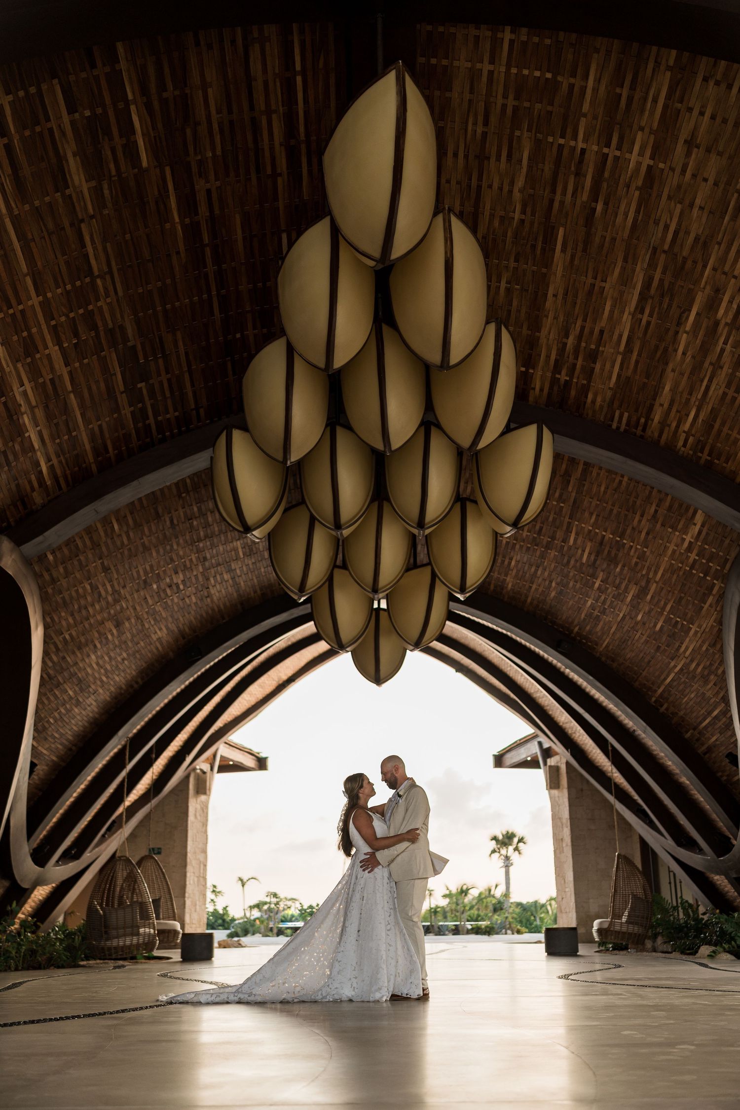 Bride and groom embrace under ornate chandelier in elegant open-air wedding venue with arched wooden ceiling.