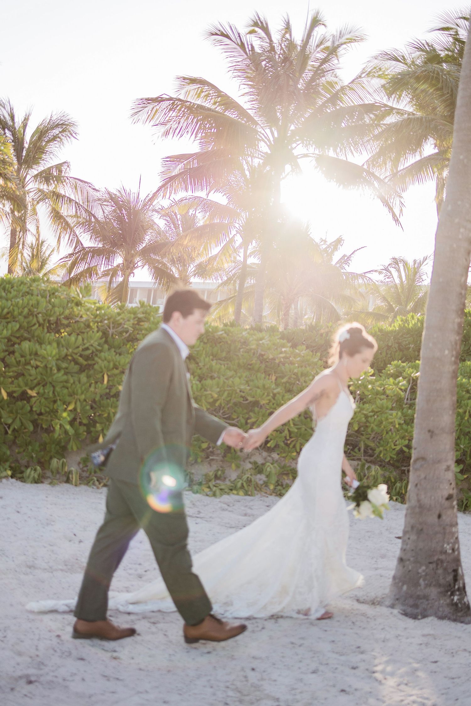 Bride and groom walking hand in hand on a sunny beach with palm trees, bride in white gown, groom in gray suit.