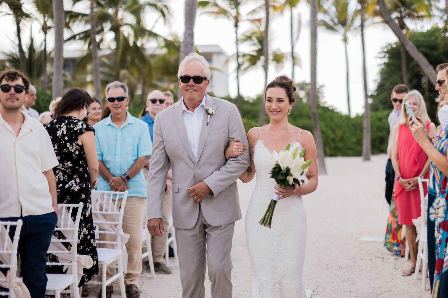 Bride walks down beach aisle with escort, holding white bouquet, guests seated in white chairs, palm trees backdrop.