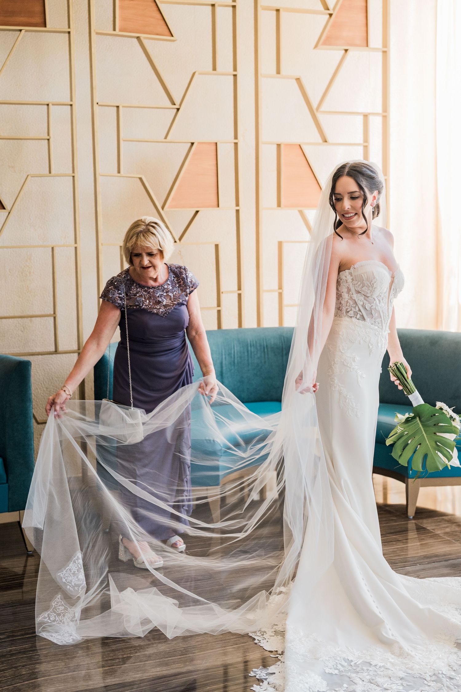 Bride in lace gown holds bouquet while mom arranges flowing veil in elegant hotel suite. Puerto Vallarta wedding photographer