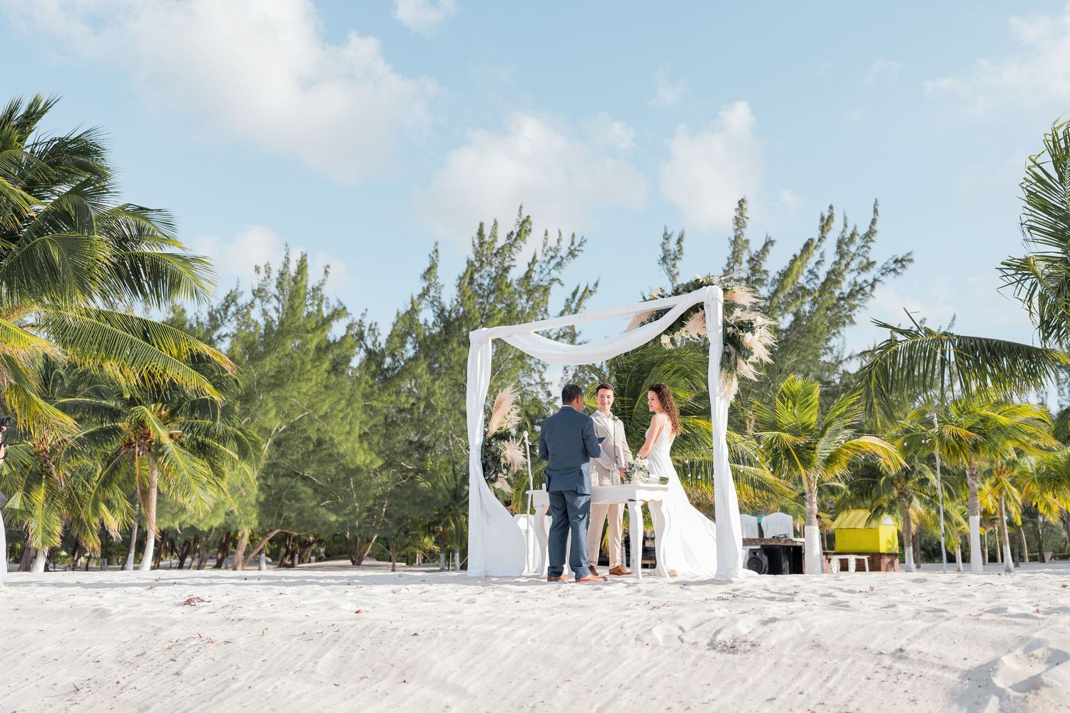 Couple exchanging vows at a beachfront elopement ceremony under a white arch surrounded by tropical palm trees.