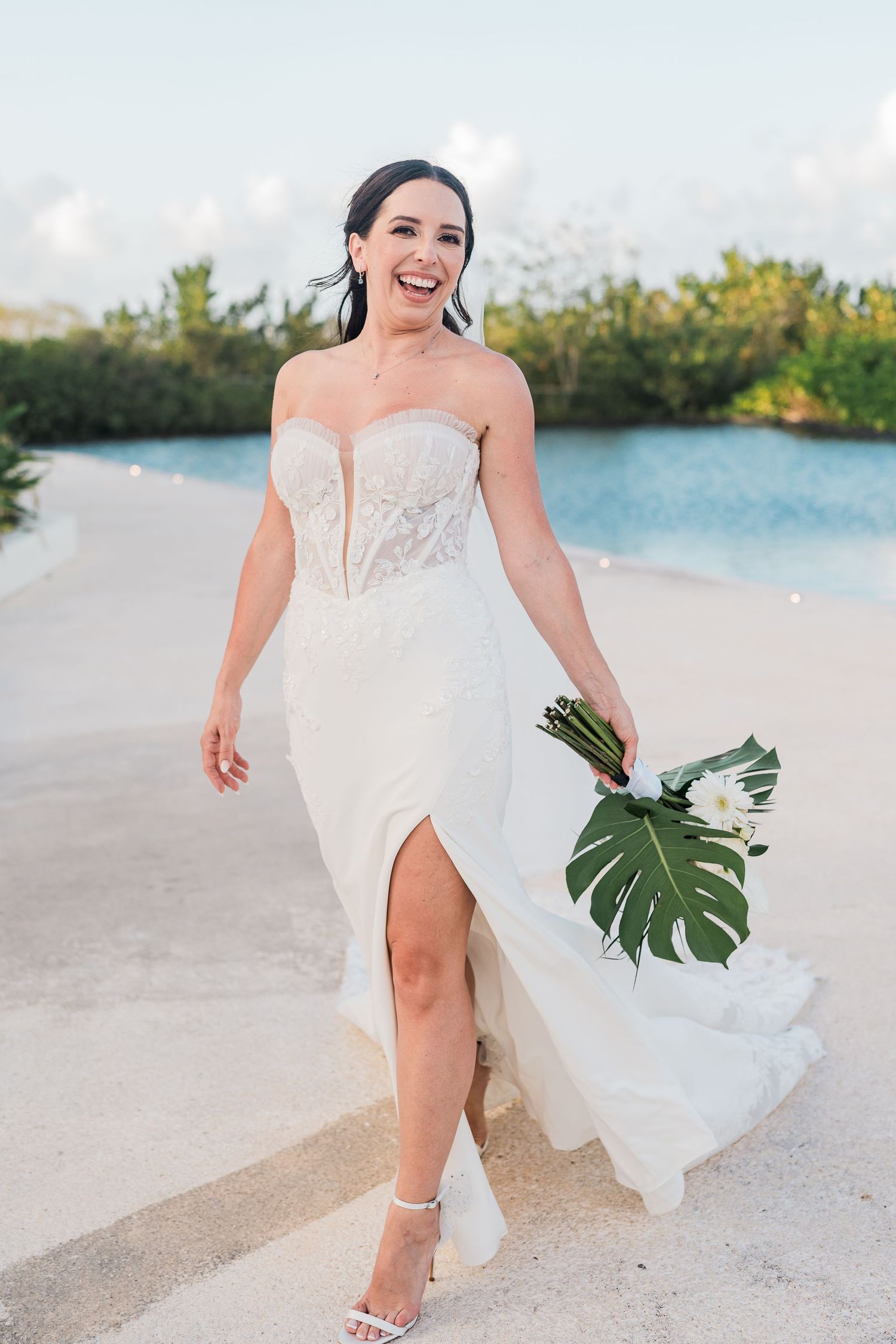 Smiling bride in strapless lace wedding gown with slit holds tropical bouquet walking along sandy beach path.