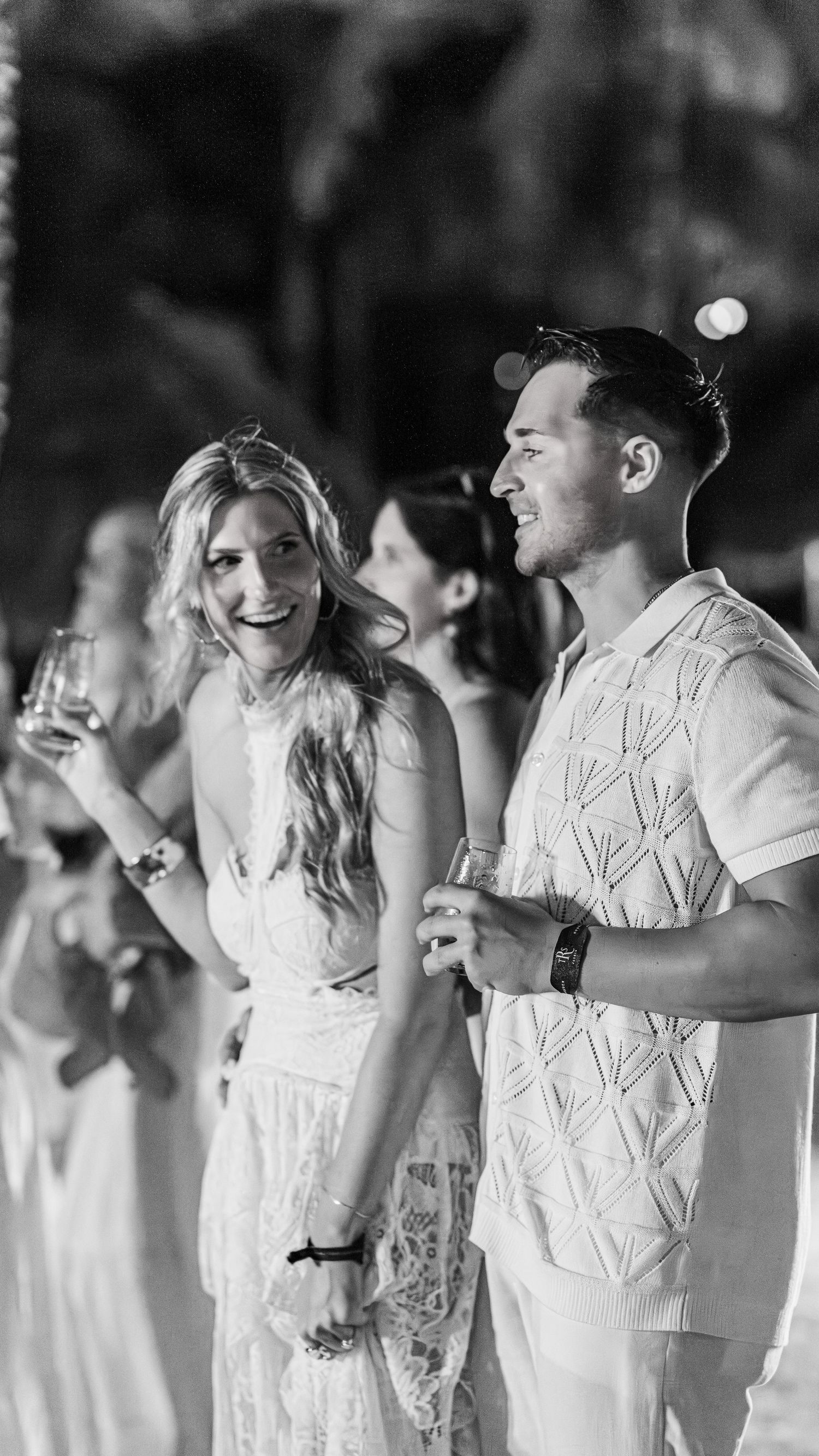 Black and white photo of a smiling couple holding drinks at an outdoor evening celebration or wedding reception.