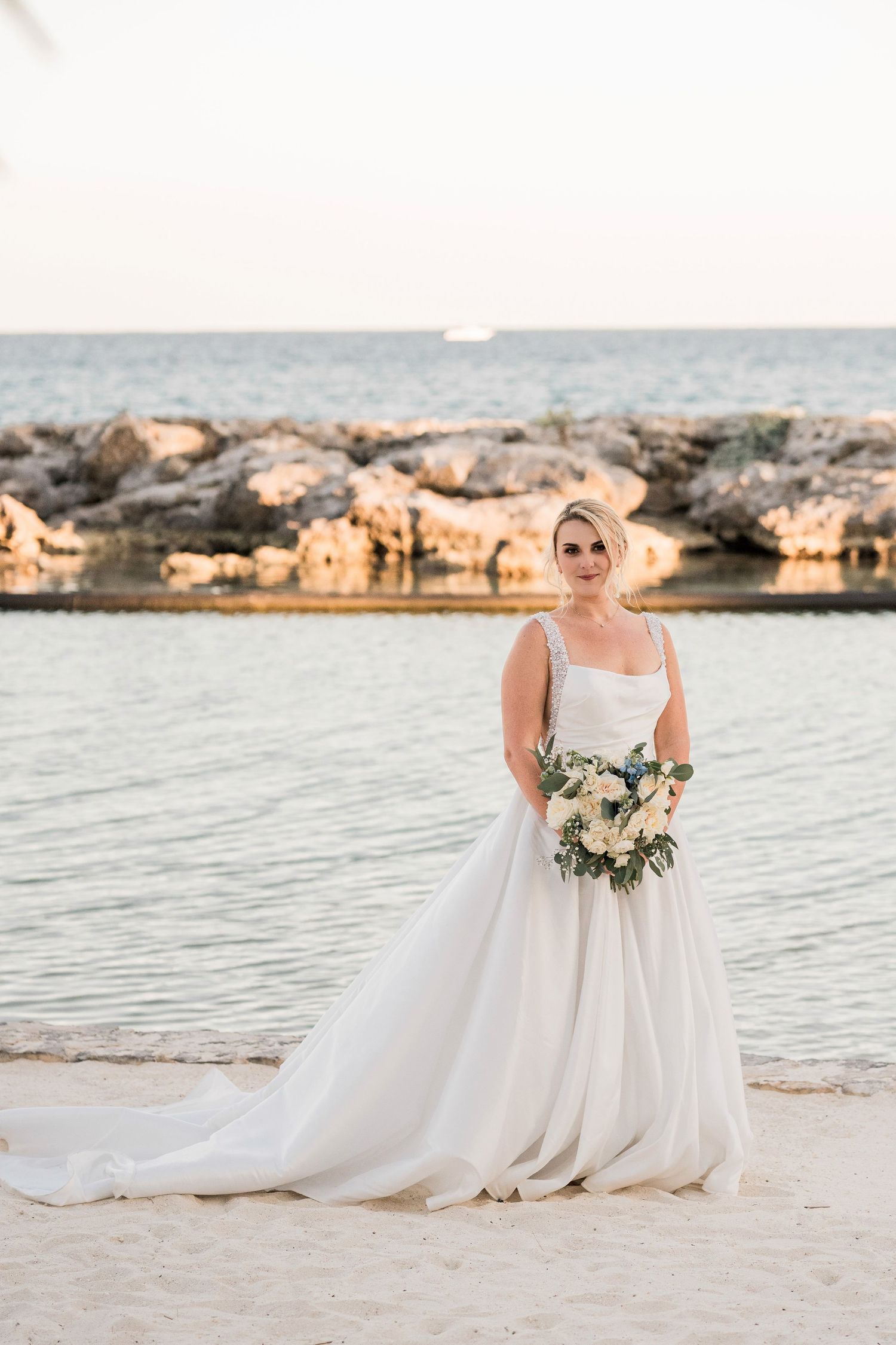 Bride in a white ball gown holding a bouquet poses on a sandy beach with rocky shoreline and ocean backdrop.