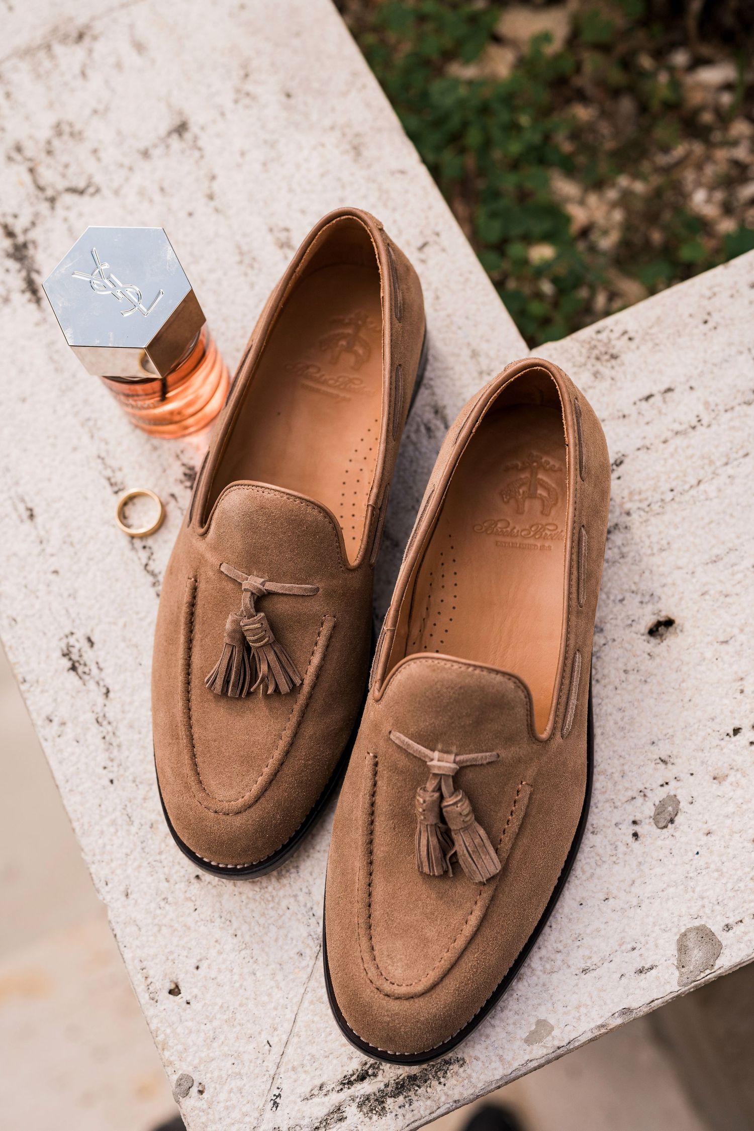 Brown suede tassel loafers displayed on a white surface beside YSL perfume and gold wedding rings.