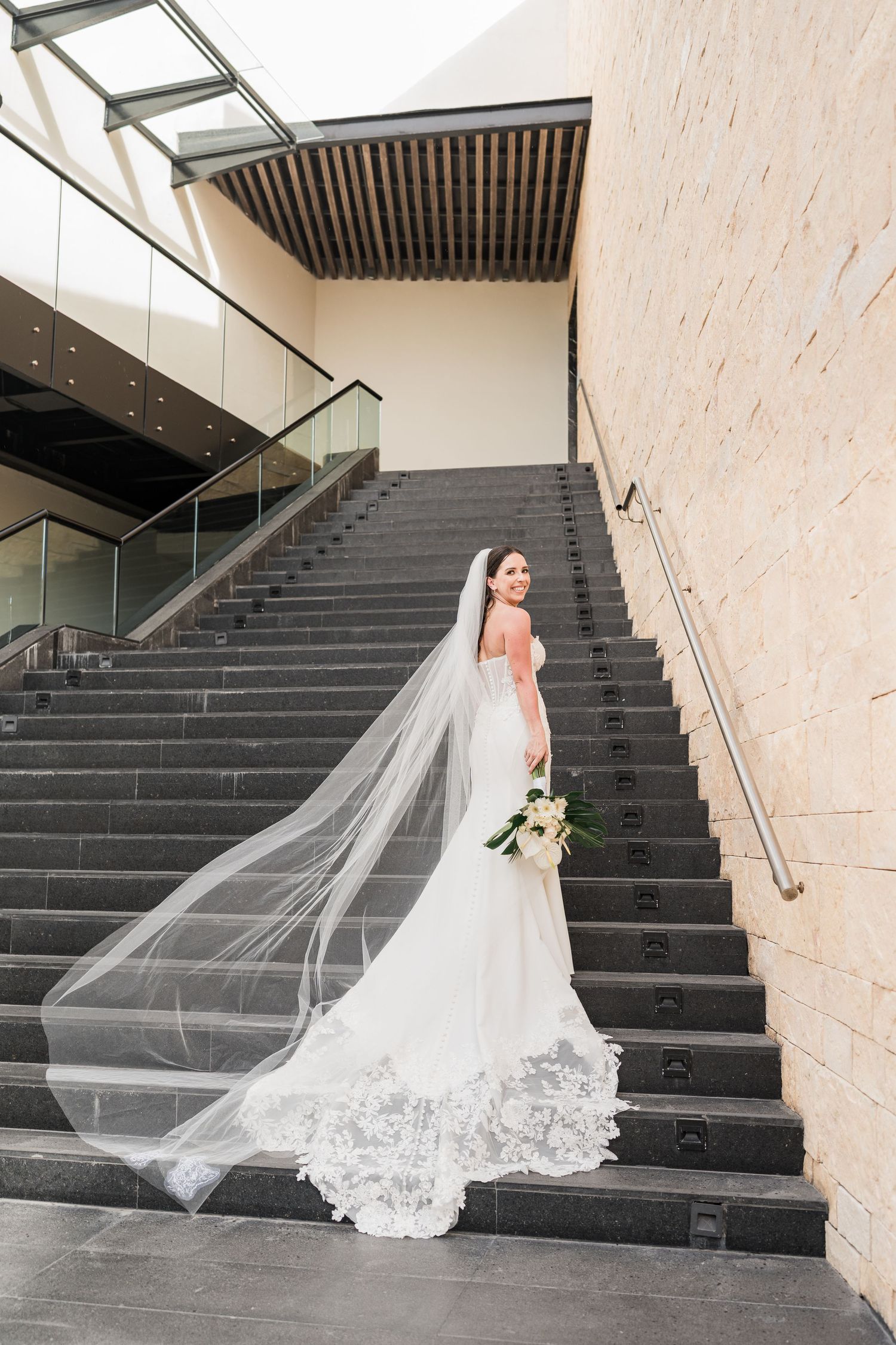 Bride in a white lace mermaid gown and cathedral veil poses on dark stone staircase in grand palladium costa mujeres