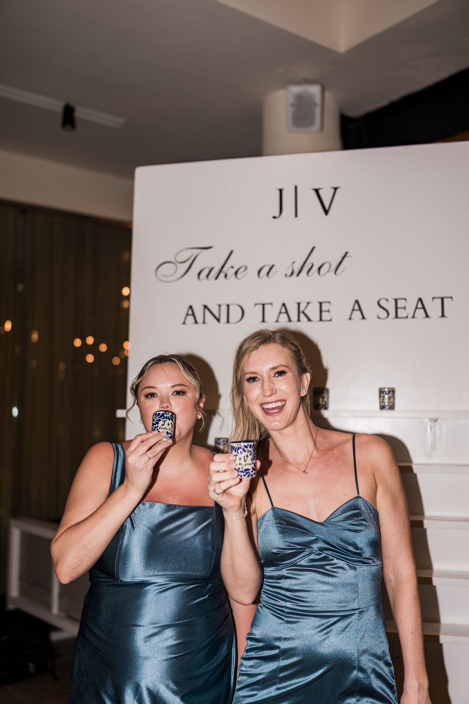 Two bridesmaids in blue satin dresses taking shots at a wedding reception near a sign reading Take a Shot and Take a Seat.
