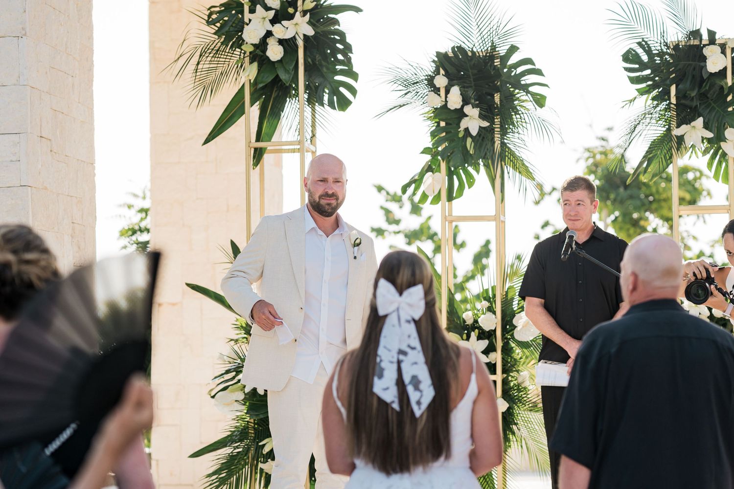 Groom in white suit stands at tropical floral wedding altar awaiting bride during outdoor ceremony.