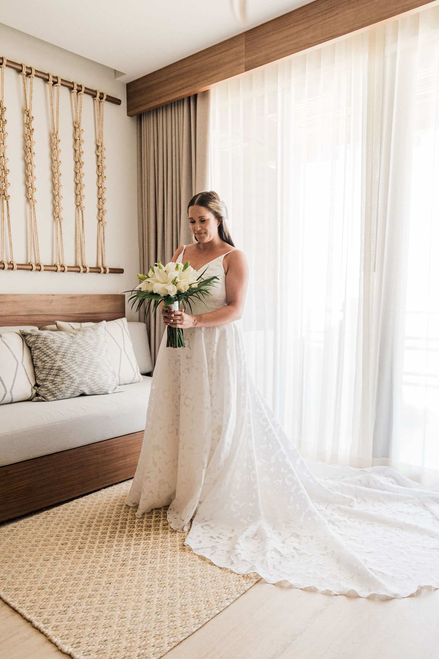 Bride in white lace gown holding bouquet of white flowers, standing by sheer curtains in elegant hotel room.