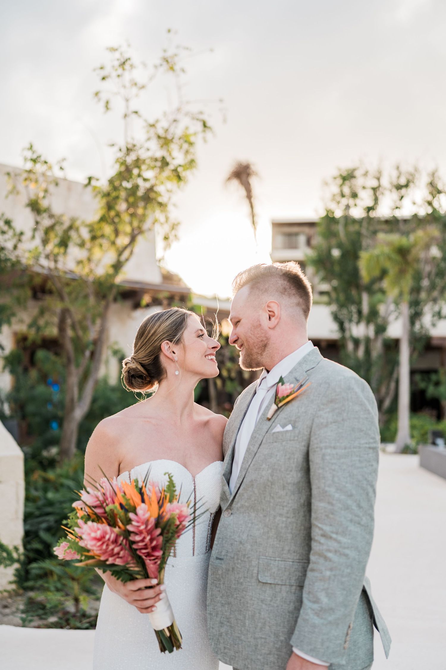 A bride and groom share a romantic moment outdoors, she holds a tropical pink bouquet, he wears a light gray suit.