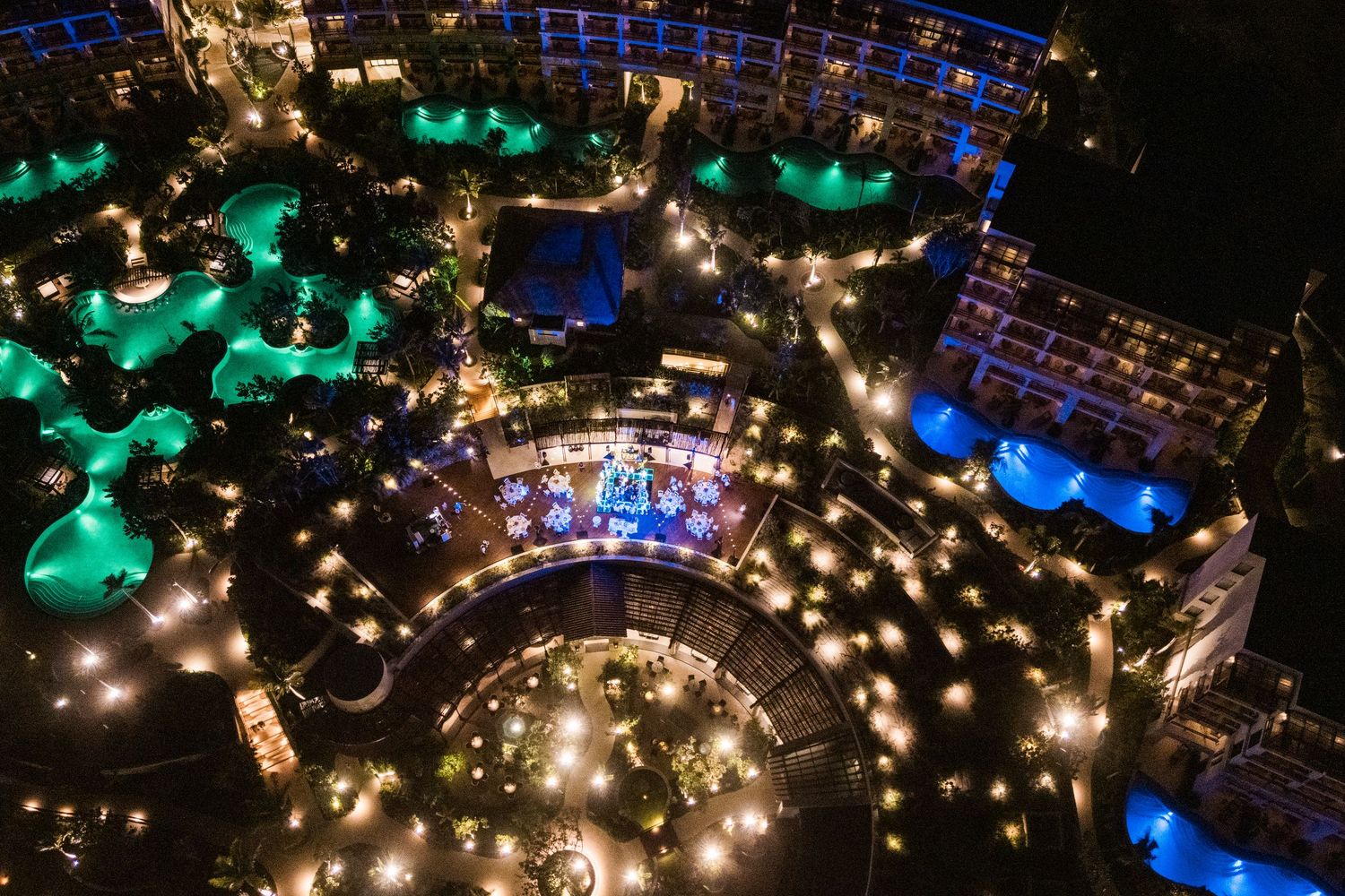Aerial night view of a luxury resort with glowing turquoise pools, string lights, and lush tropical landscaping.