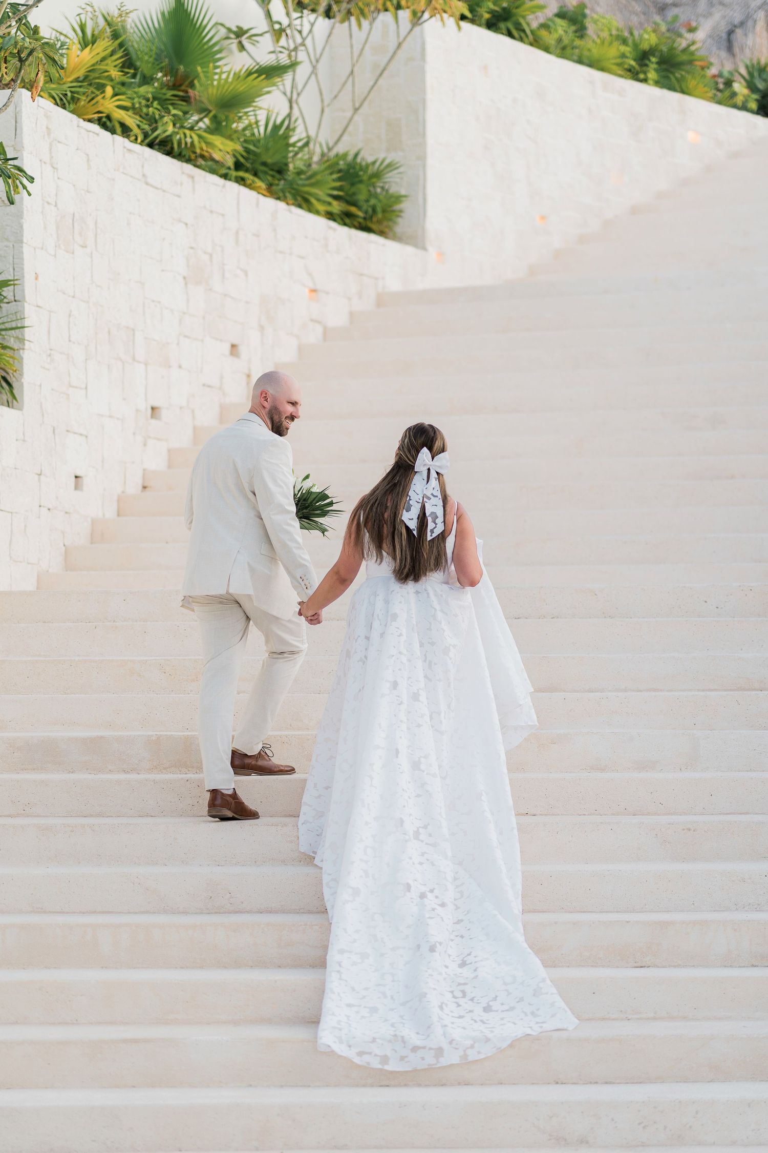 Bride in lace gown and groom in beige suit hold hands ascending white stone stairs at outdoor wedding venue.